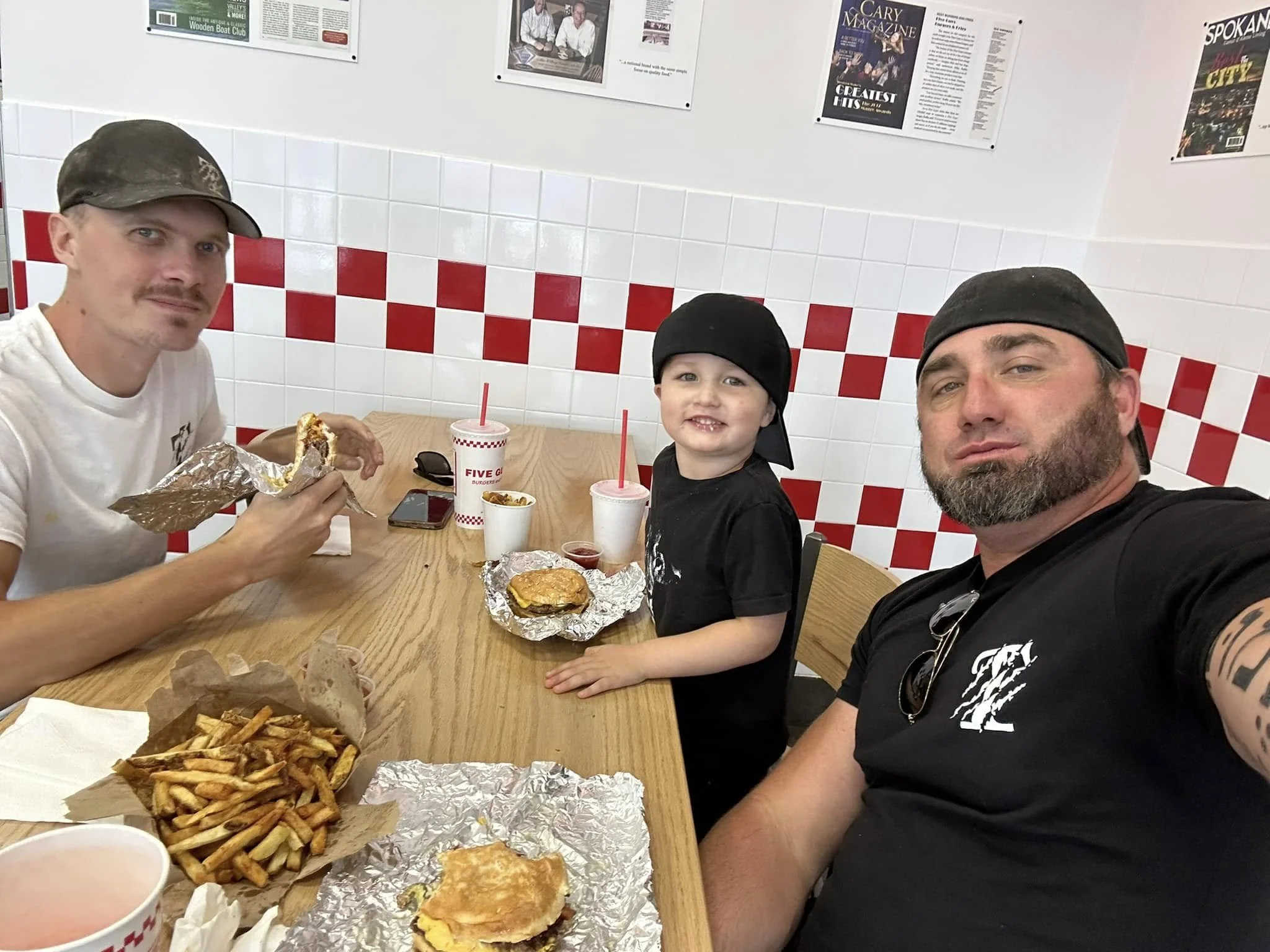 Three people sitting at a table in a fast-food restaurant, eating burgers and fries. The adults are wearing black shirts; the child is smiling. Fast food cups and foil wrappers are on the table.