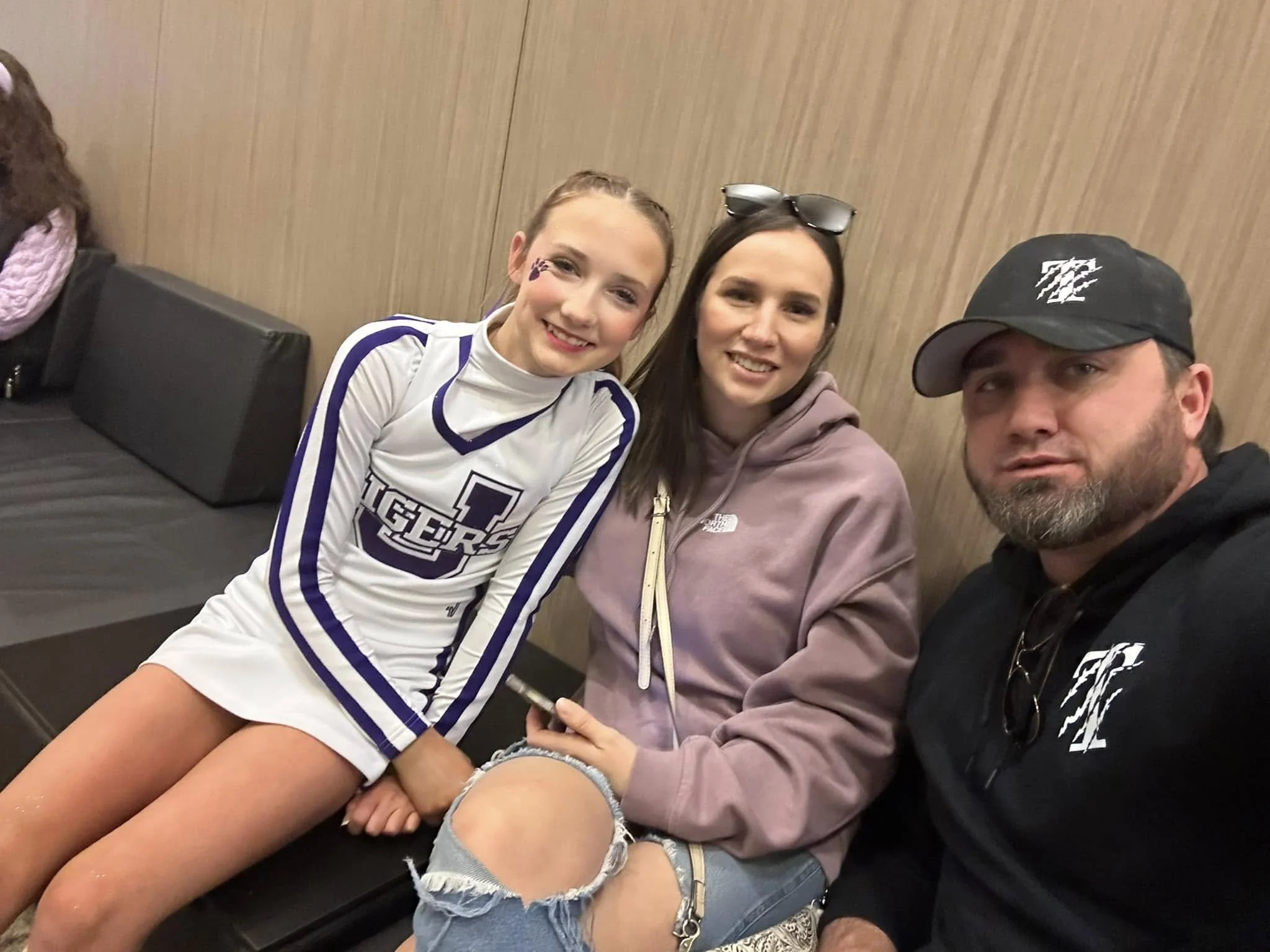 A young girl in a white and purple cheerleading uniform sits on a bench next to two adults, a woman in a pink hoodie and a man in a black hoodie and cap.