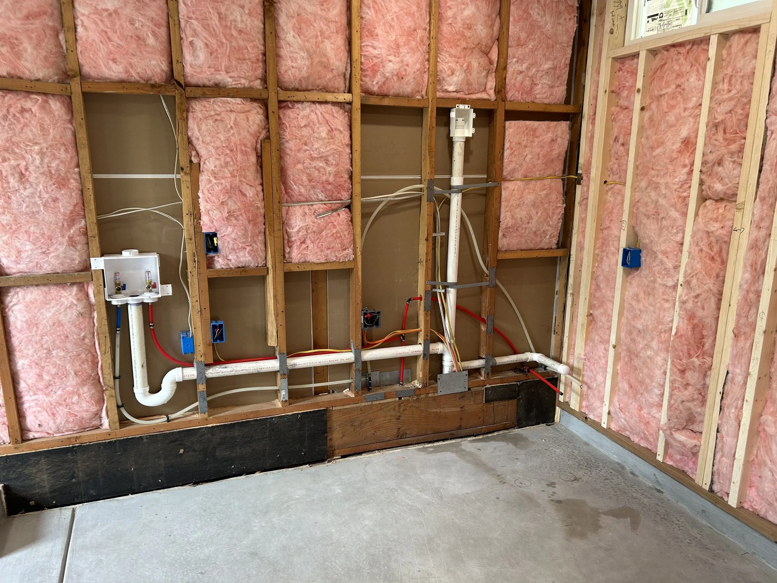 Exposed interior wall with pink insulation, electrical wiring, and plumbing pipes in a construction site.