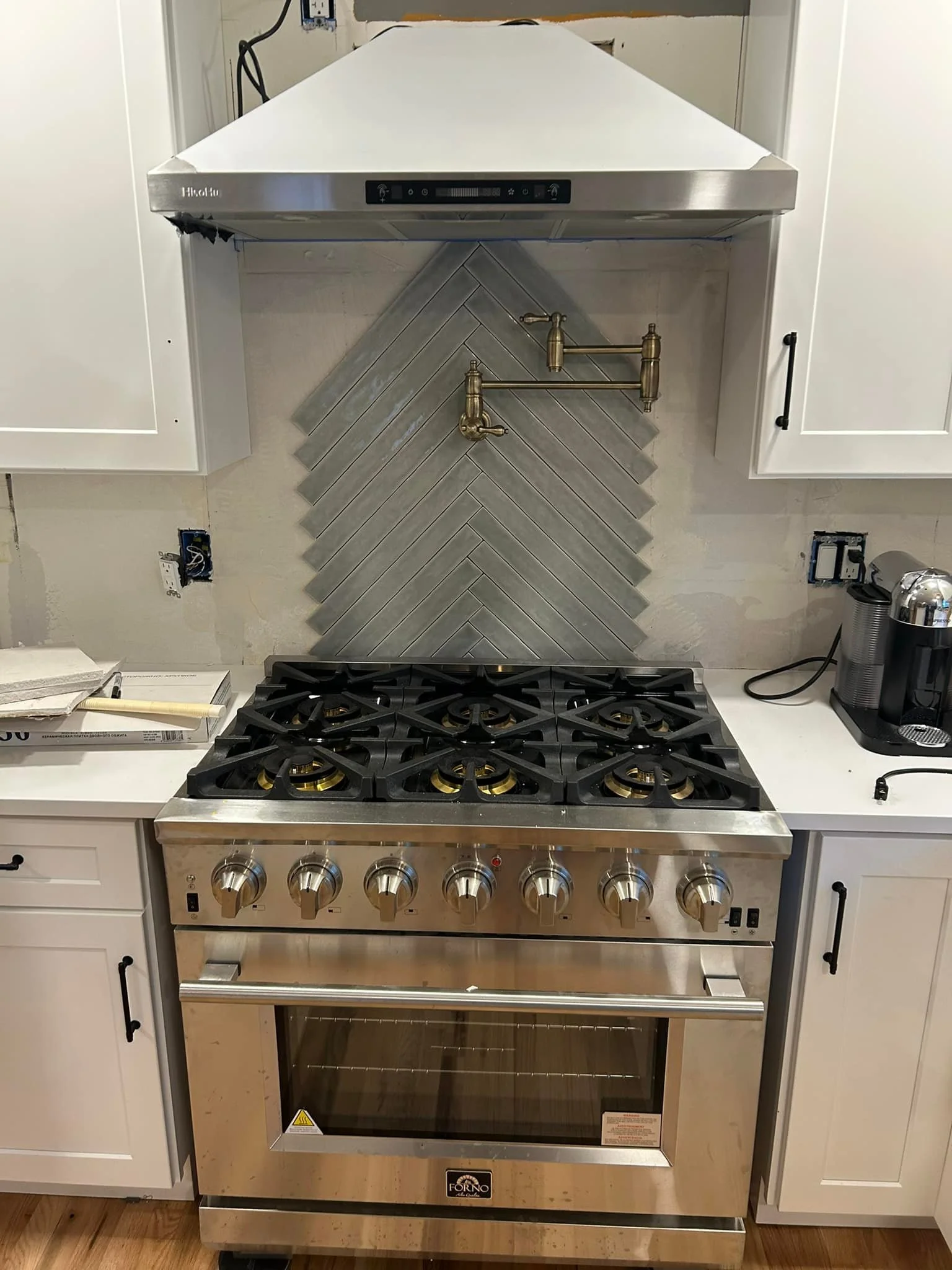 Stainless steel gas stove with six burners, stainless steel range hood, white cabinetry, gray herringbone tile backsplash, and a brass pot filler above the stove.