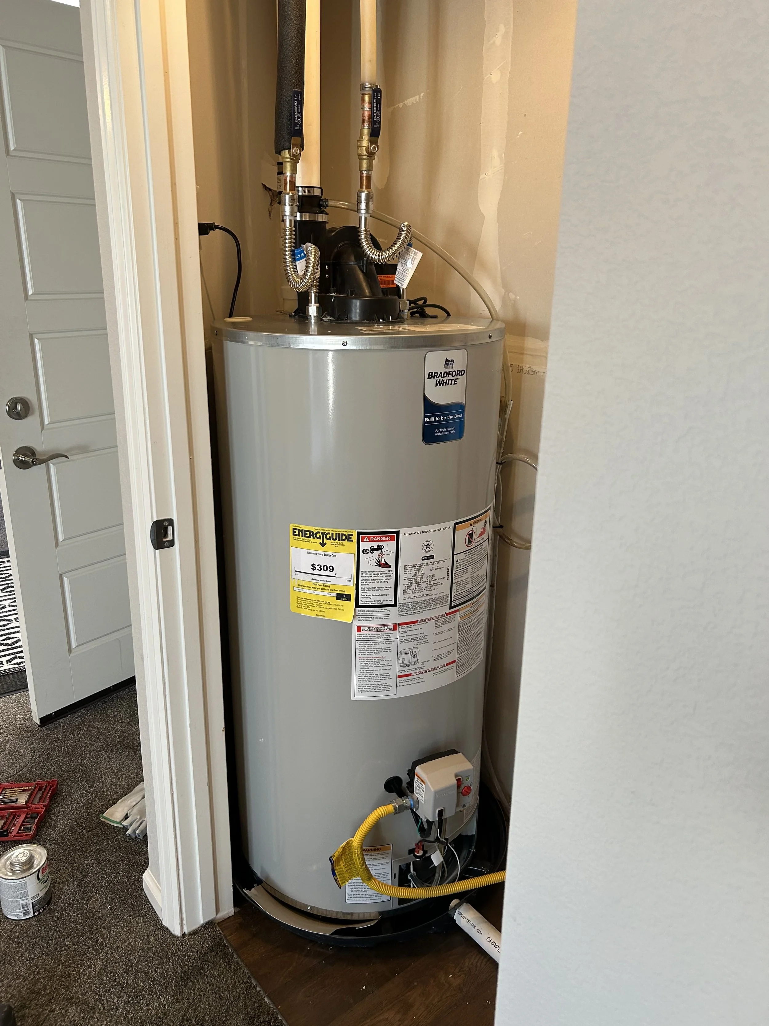 Residential water heater installed in a utility closet with attached pipes and labels.
