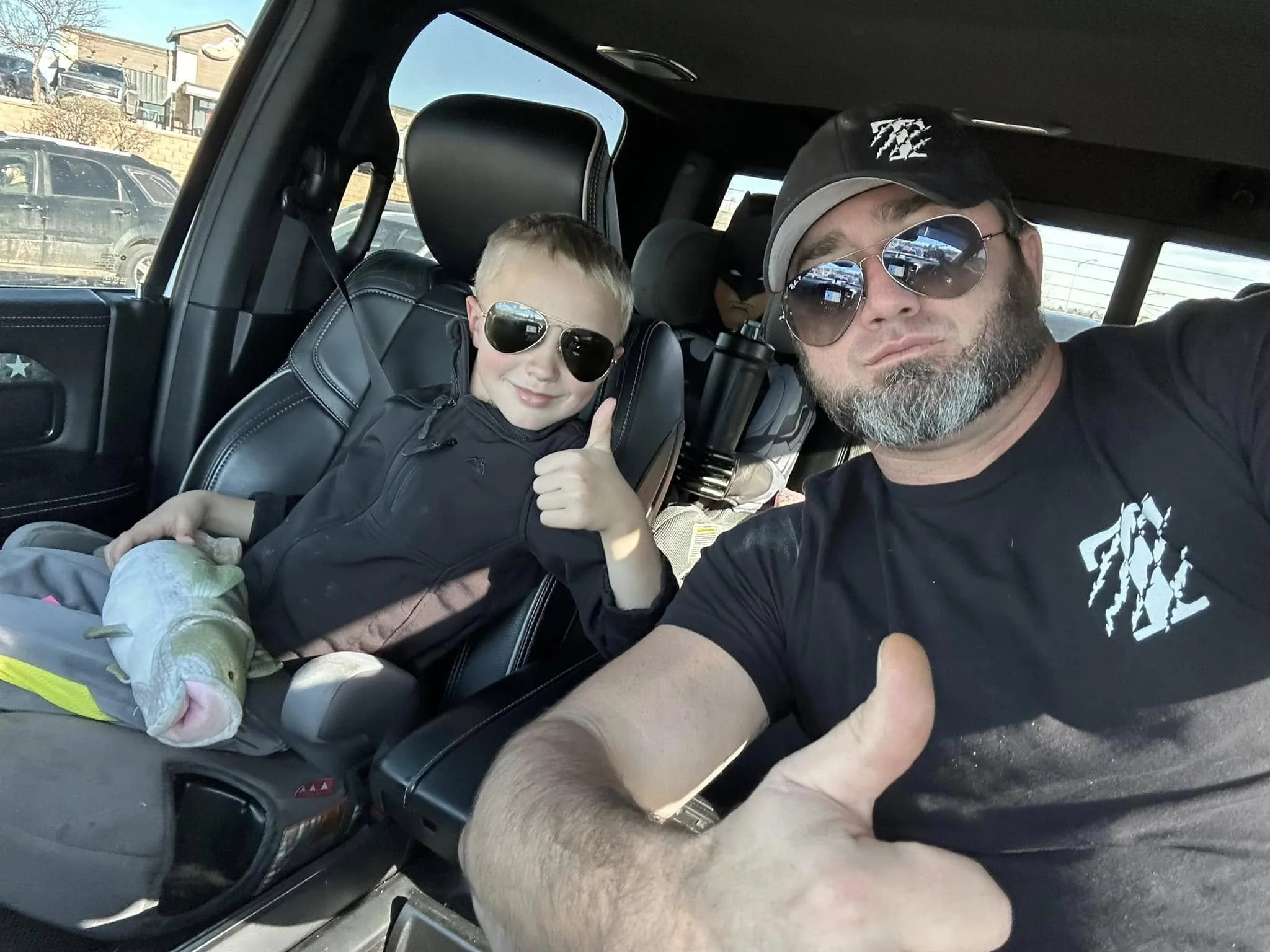 A man and a child wearing sunglasses give thumbs-up while sitting in a car. The child is holding a toy fish. The man is wearing a black cap and T-shirt with a white design.