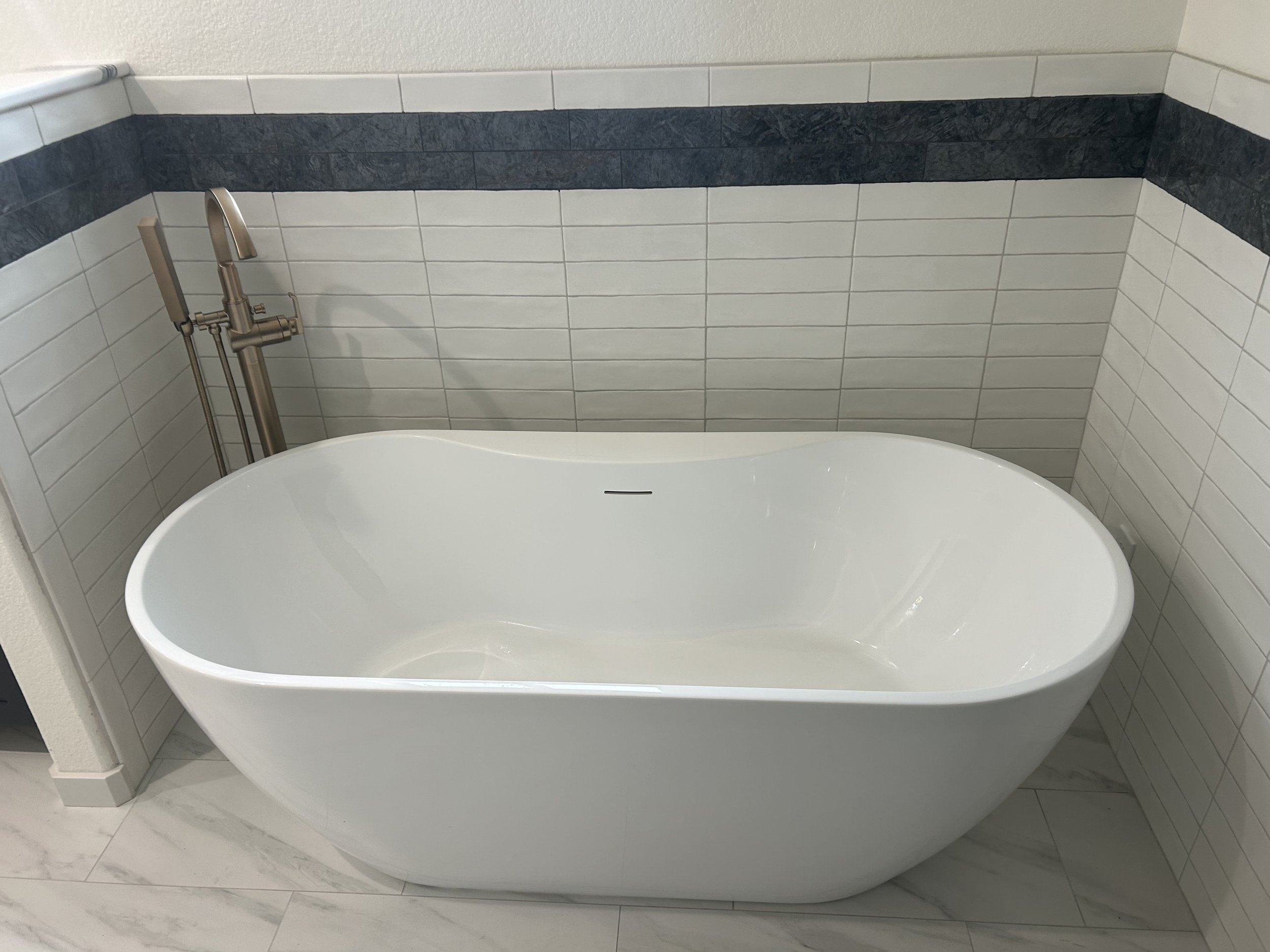 A modern white freestanding bathtub with a sleek faucet, set against a tiled wall with horizontal gray and white tiles.