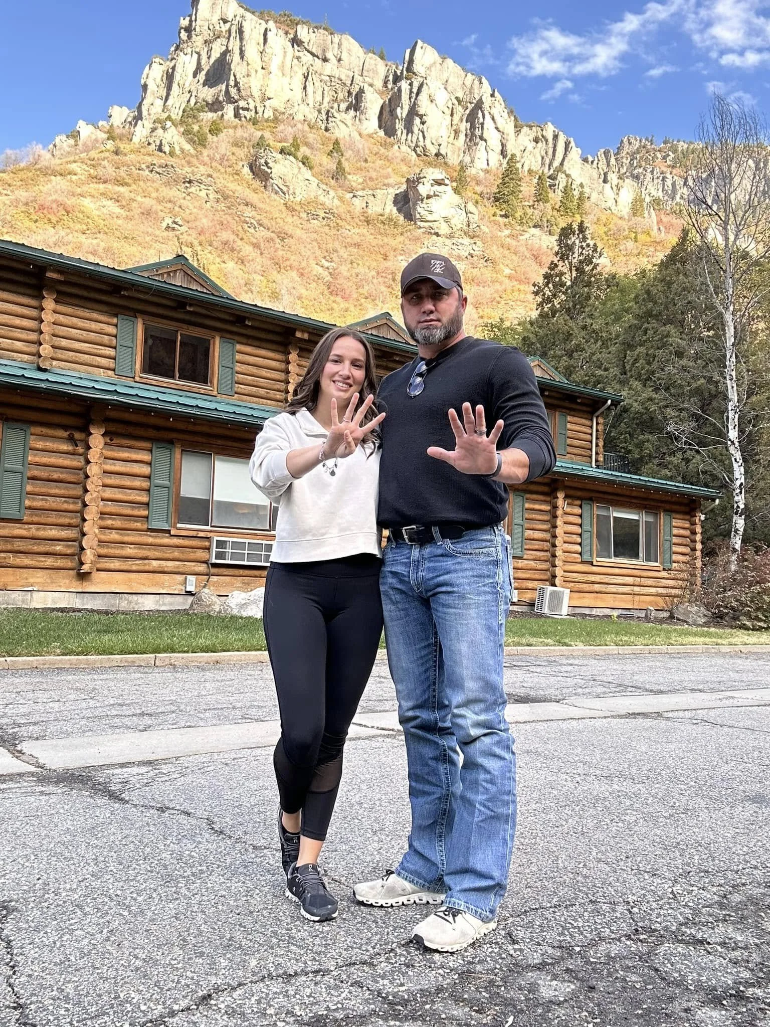 Two people standing in front of a log cabin with a rocky hill in the background, both smiling and holding their hands up. One person is wearing a black long-sleeve shirt and blue jeans, and the other is in a white sweater and black leggings. Clear sk