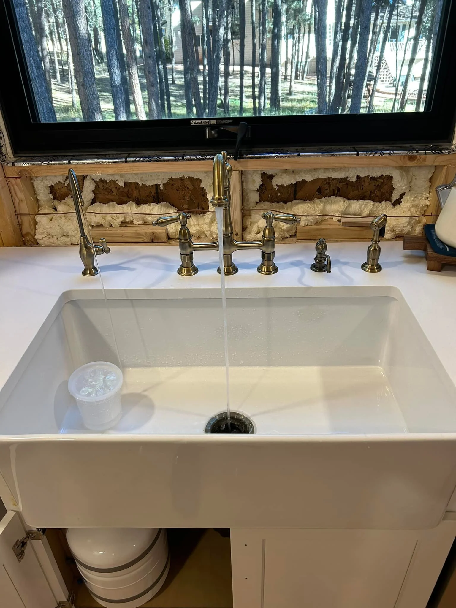 A white kitchen sink with water running from a brass faucet, surrounded by construction materials and insulation in progress. A clear container is in the sink. A window with a view of trees is above the sink.