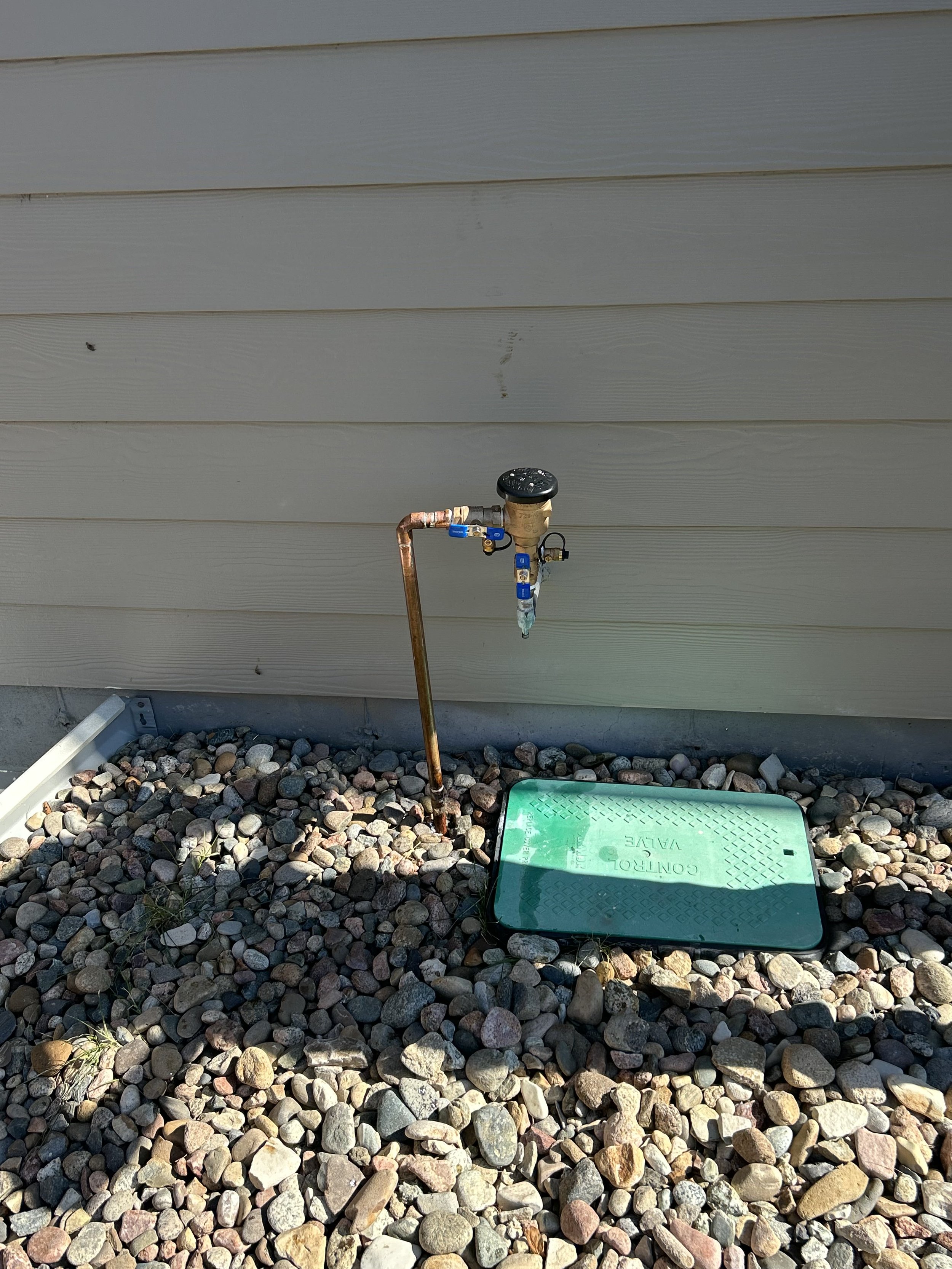 A water meter and valve setup near a building wall, surrounded by gravel. The water meter is positioned above a copper pipe with shut-off valves. A green control valve cover is situated on the ground next to the setup.