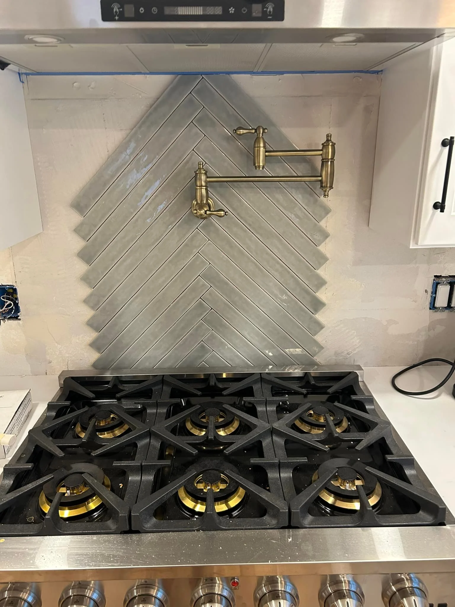 Kitchen stovetop with six burners and a herringbone tile backsplash, featuring a brass pot filler faucet.