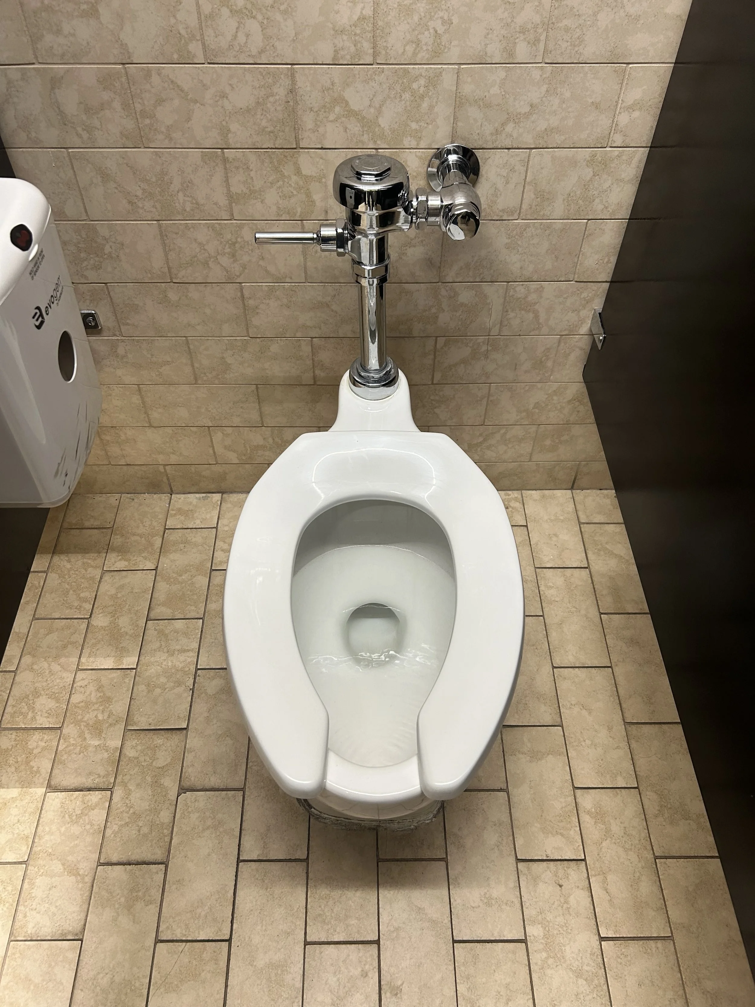 Public restroom toilet with a flushometer and tiled wall and floor.