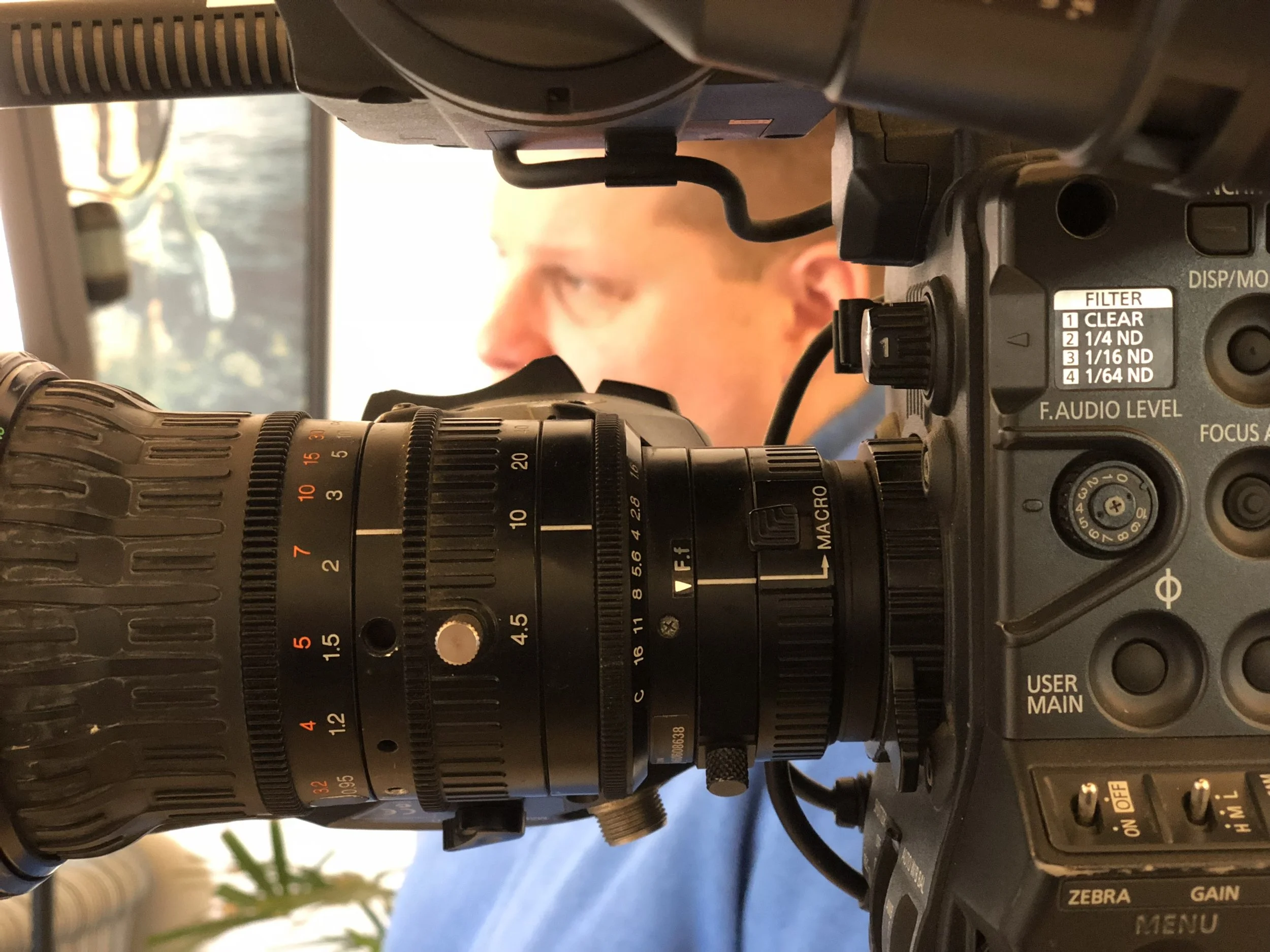 Close-up of a camera with macro lens, focus controls, and filter settings, partially revealing a person in the background.