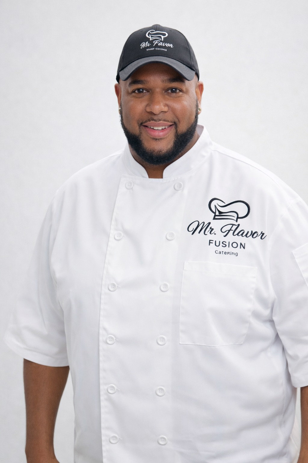 A man smiling wearing a white chef uniform with 'Mr. Flavor' logo and a black cap with the same logo.