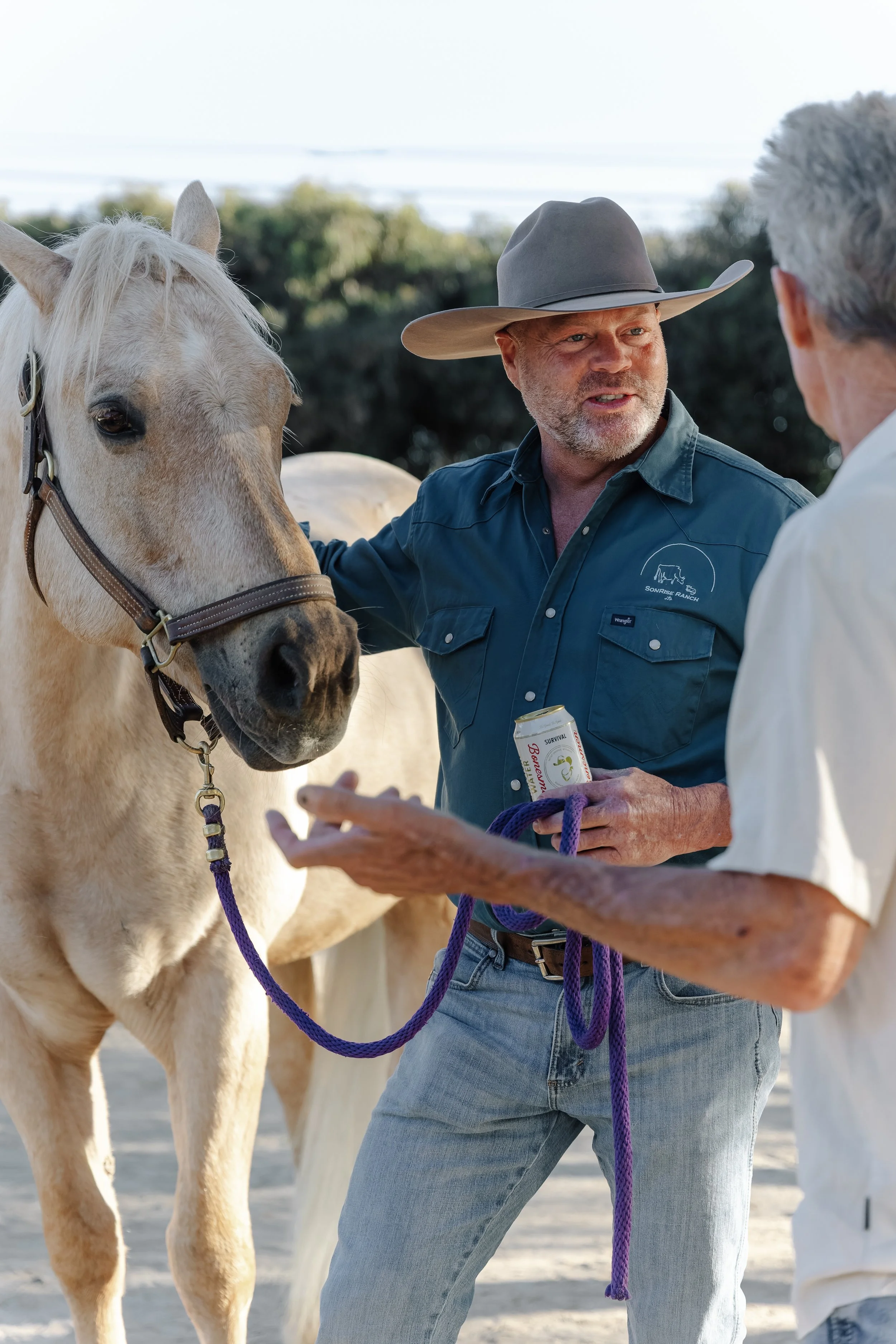 Two men talking near a light-colored horse with a purple lead rope in a rural outdoor setting.
