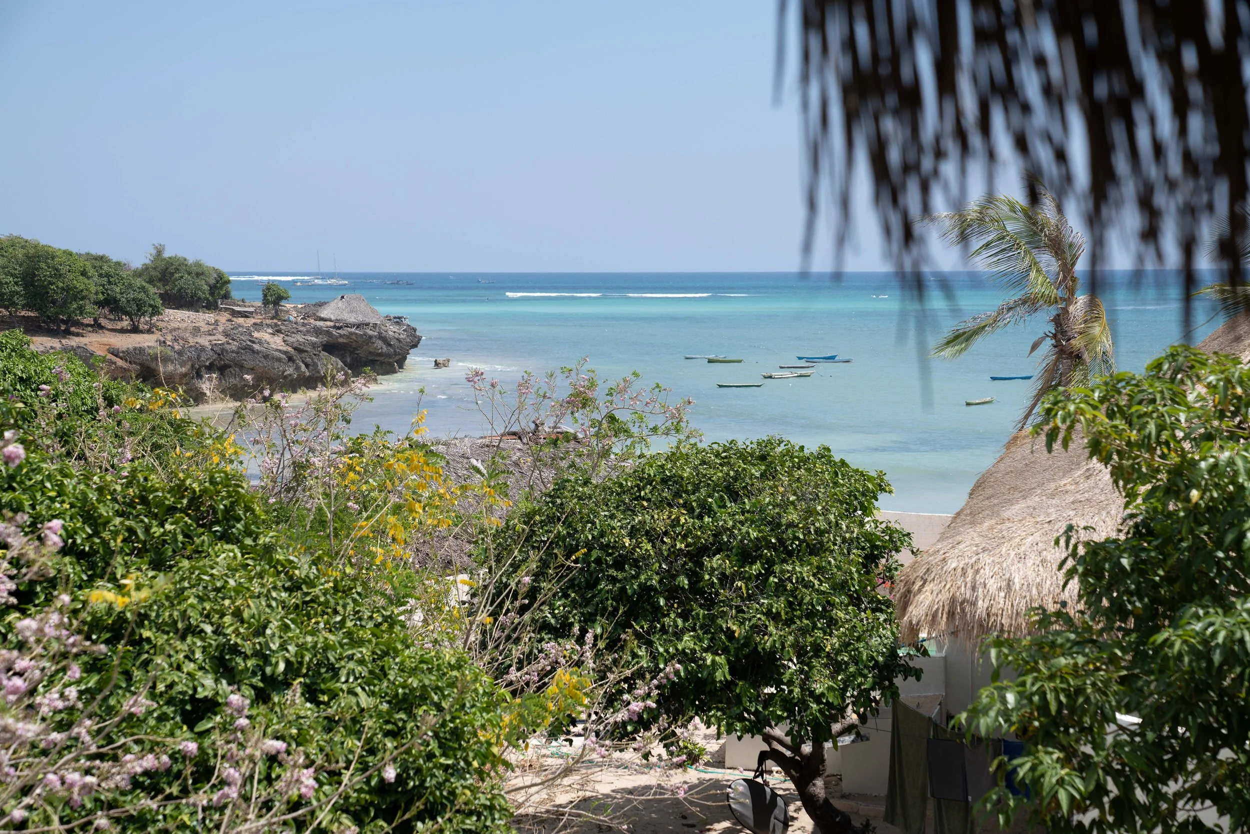 Tropical beach with turquoise water, small boats, lush greenery, rocky shoreline, and a thatched roof structure.