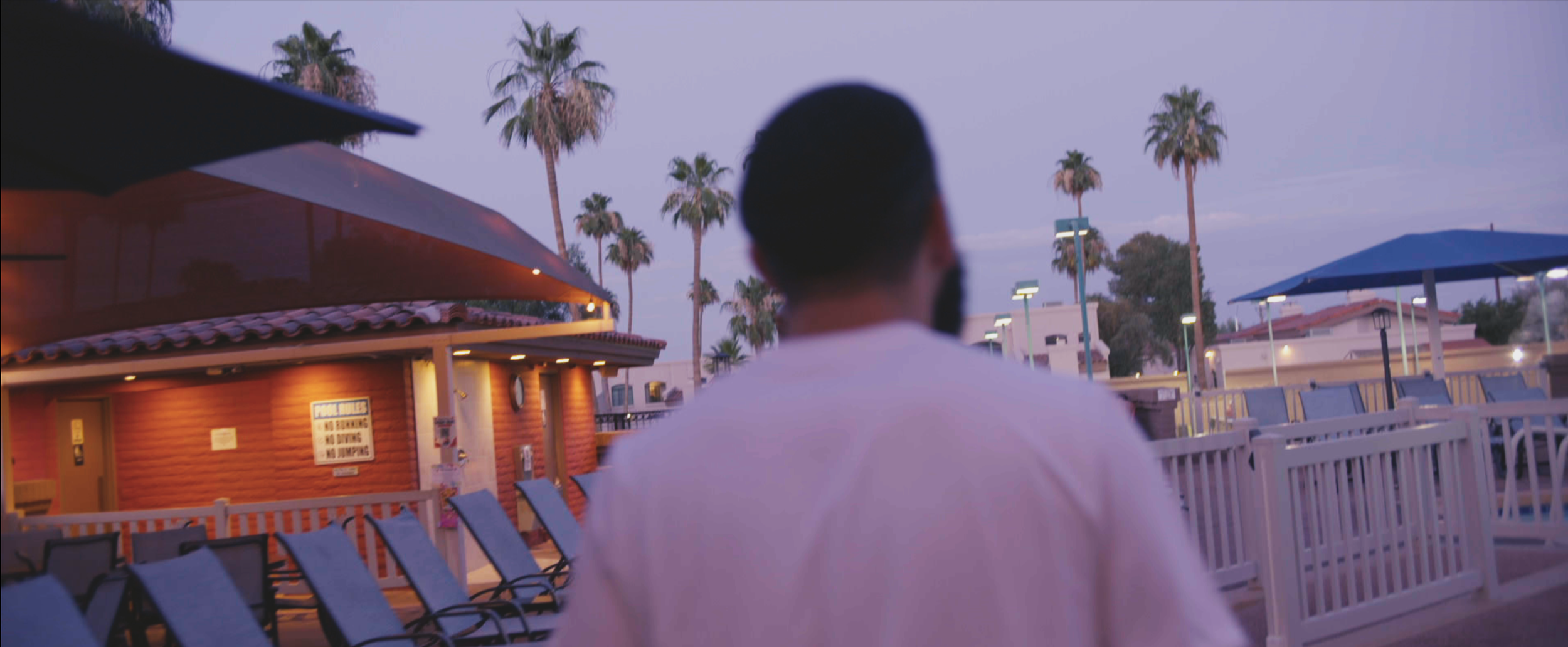 A man with short dark hair and a beard, seen from behind, looking at an outdoor swimming pool area during dusk, with palm trees and building structures in the background.