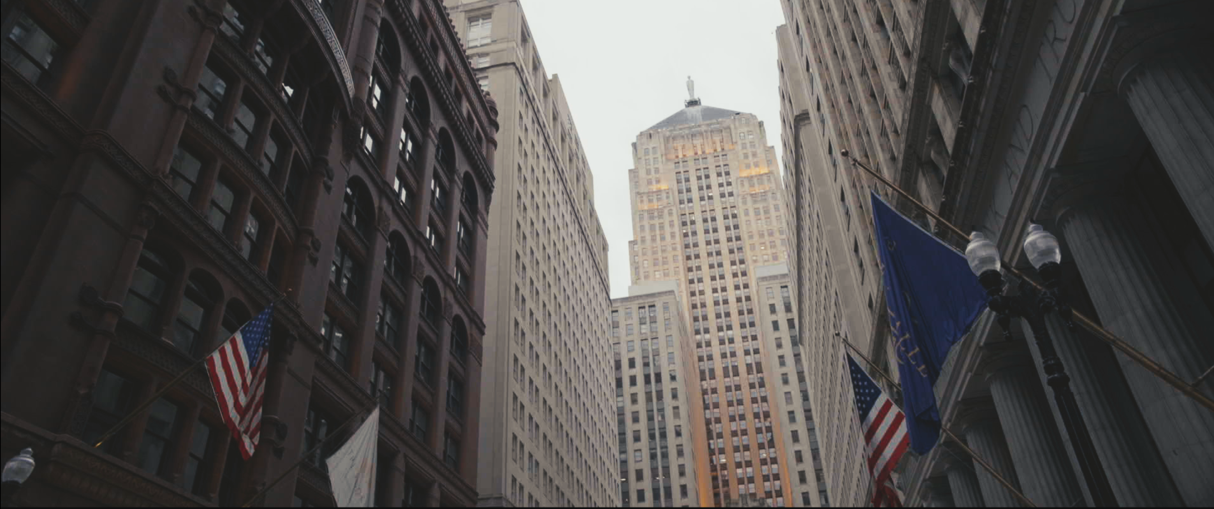 View of tall skyscrapers in a city, with American flags hanging from a building on the right and flags on the left.