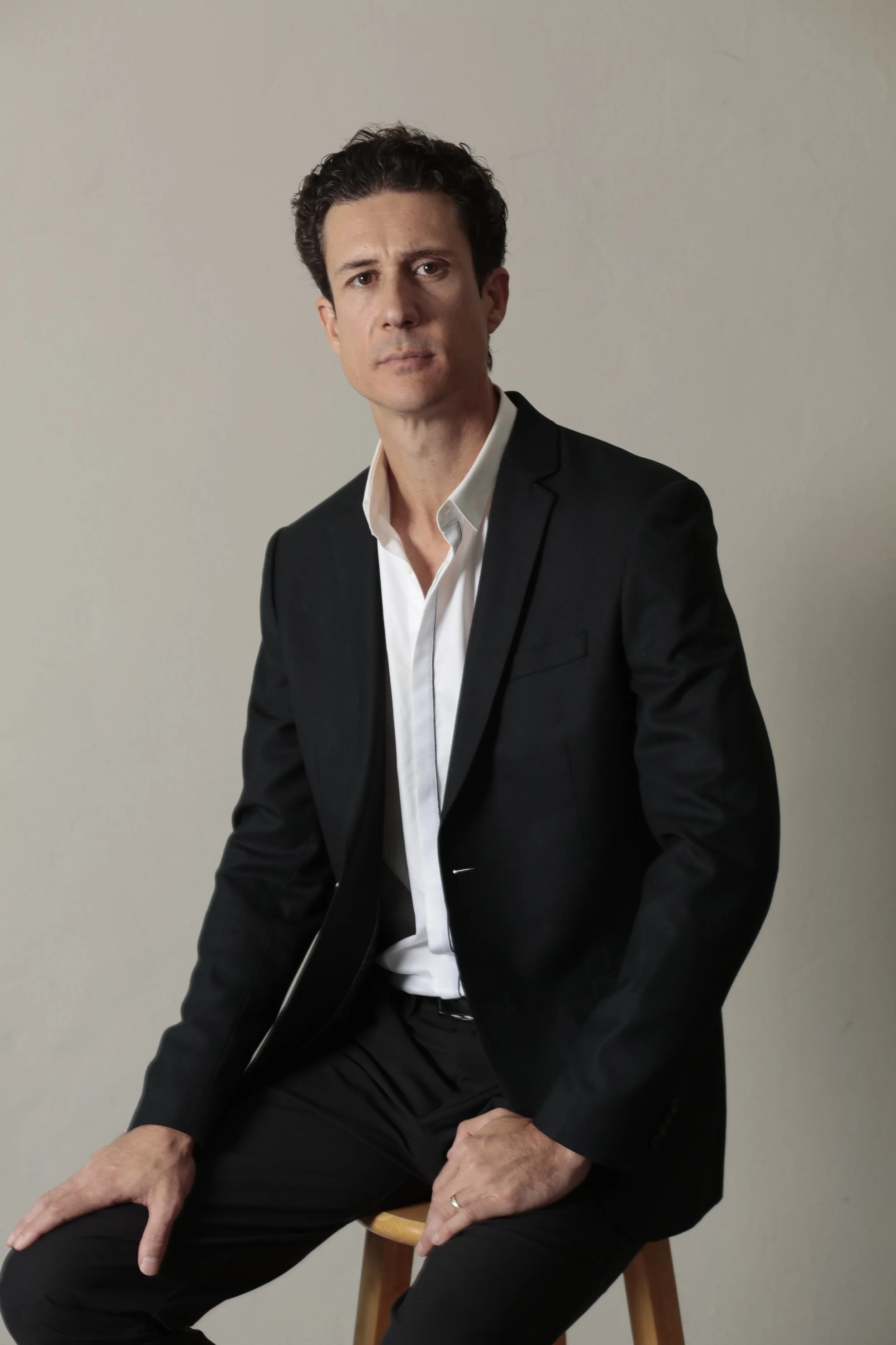 A man in a black suit with a white shirt, sitting on a wooden stool against a plain gray wall, looking directly at the camera with a serious expression.