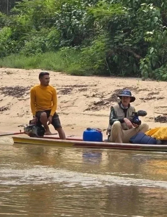 Two men on a small boat on a river, one wearing a yellow long-sleeve shirt and black shorts, the other wearing a sun hat and beige pants, holding video cameras, with a blue container and belongings in the boat, and a sandy riverbank with lush green foliage in the background.