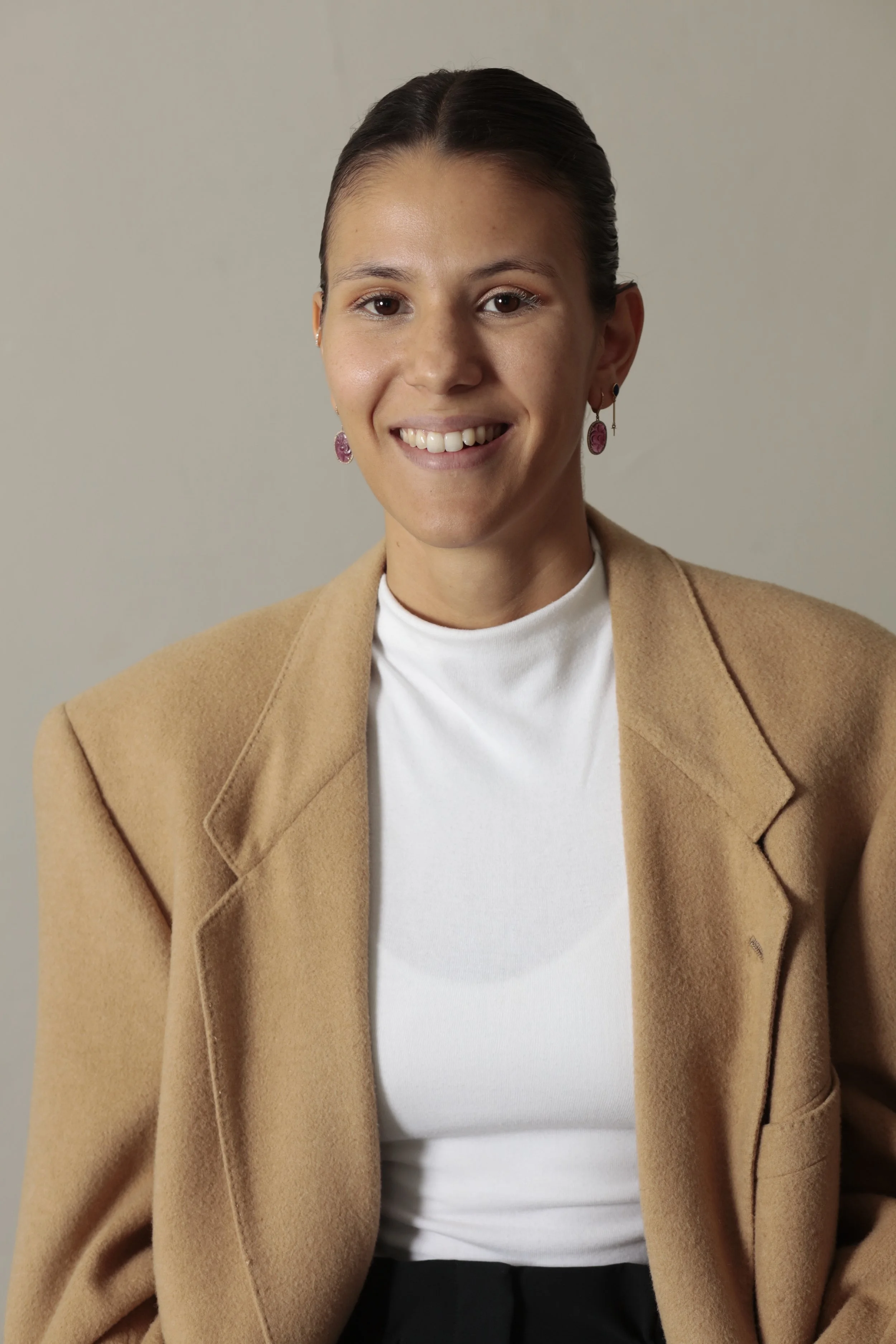 Portrait of a smiling woman with dark hair pulled back, wearing earrings, a white top, and a tan blazer against a plain background.