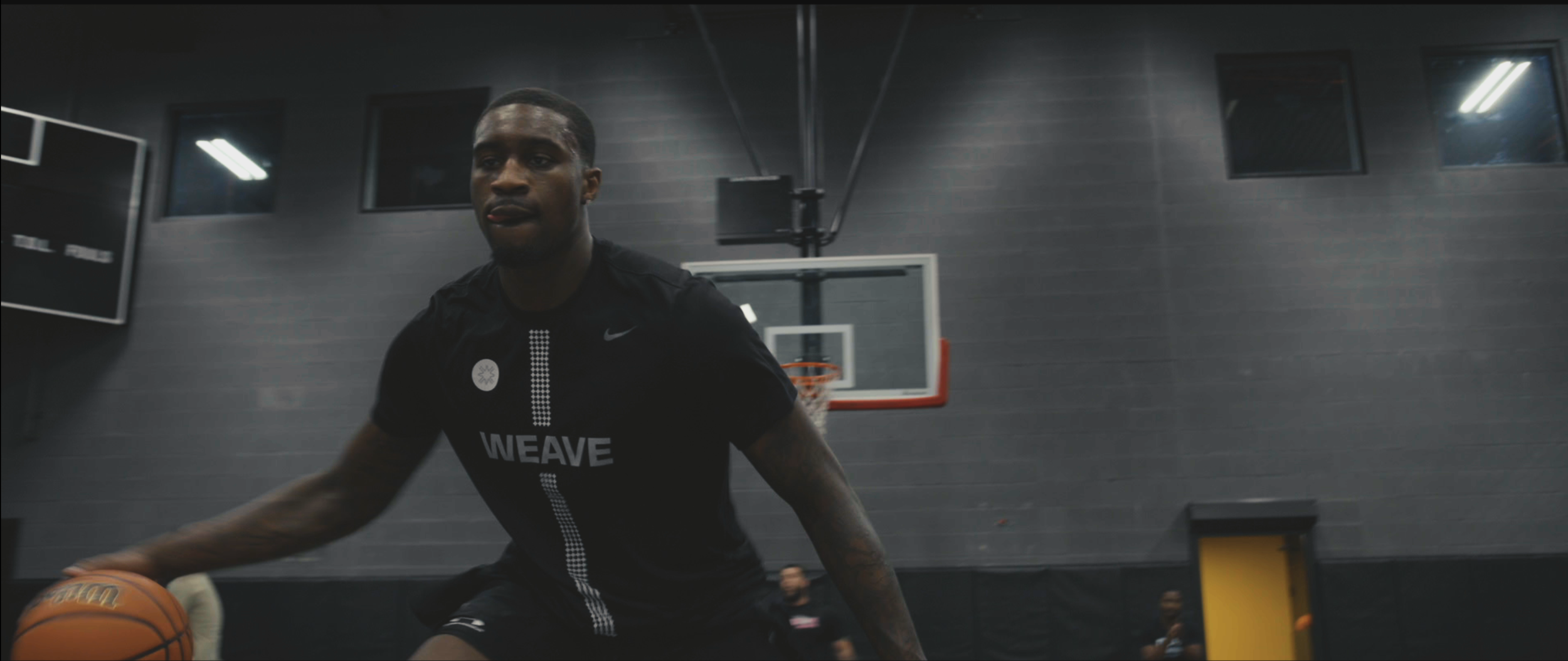A male basketball player in black sportswear dribbling a basketball on an indoor court, with a brick wall, basketball hoop, and a basketball scorer in the background.