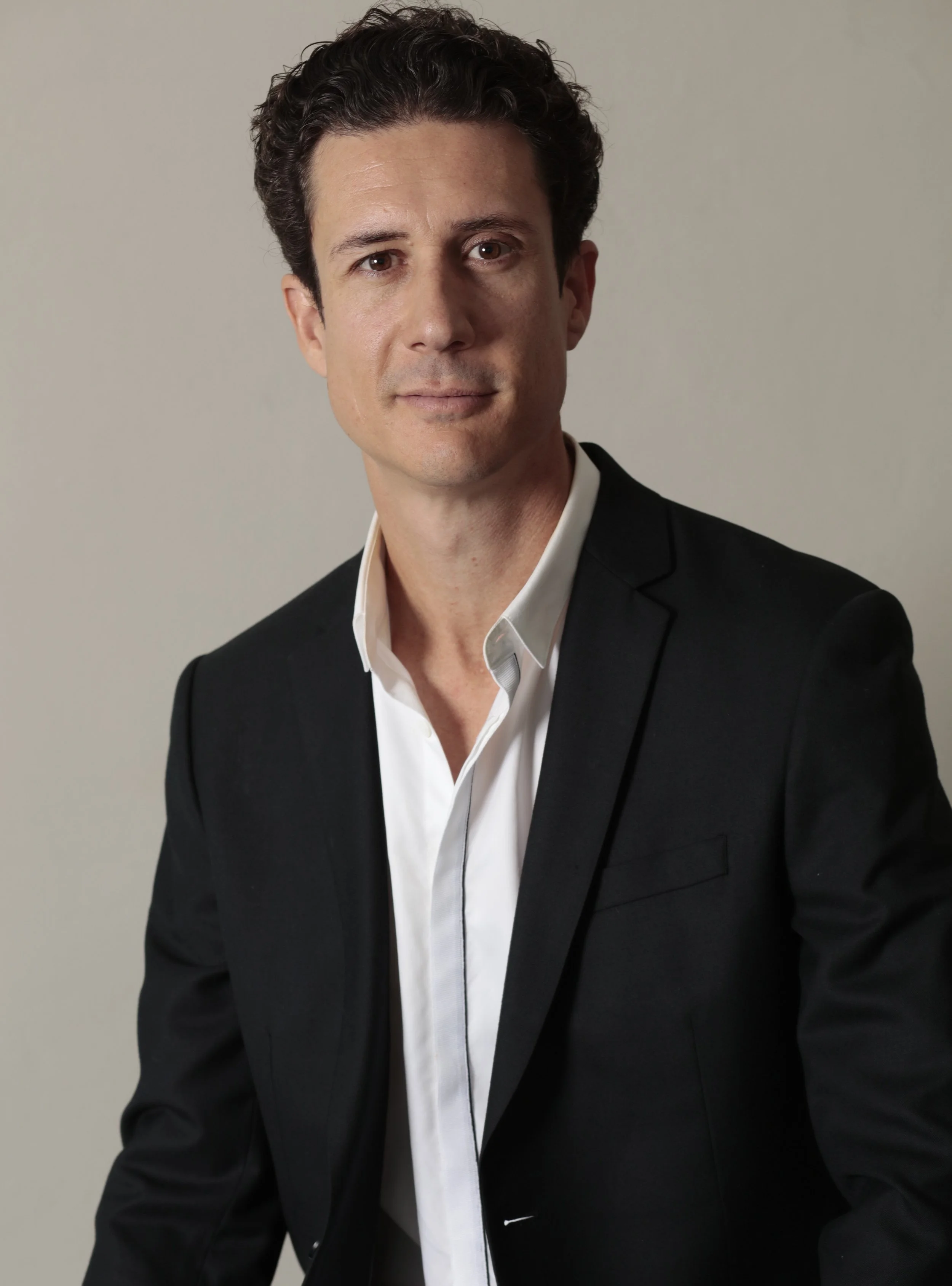 A man in a black suit with a white shirt against a plain light background.