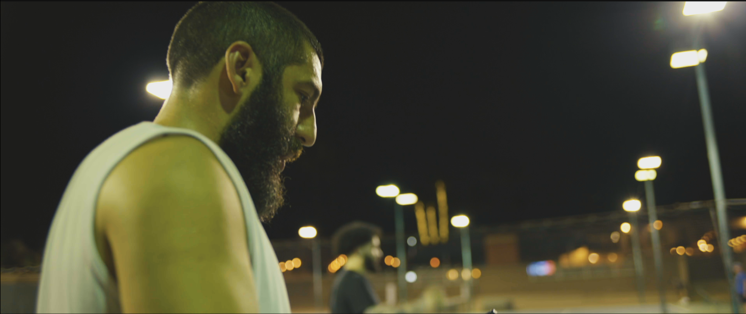 A bearded man with short hair wearing a sleeveless athletic shirt, standing outdoors at night with stadium lights in the background.