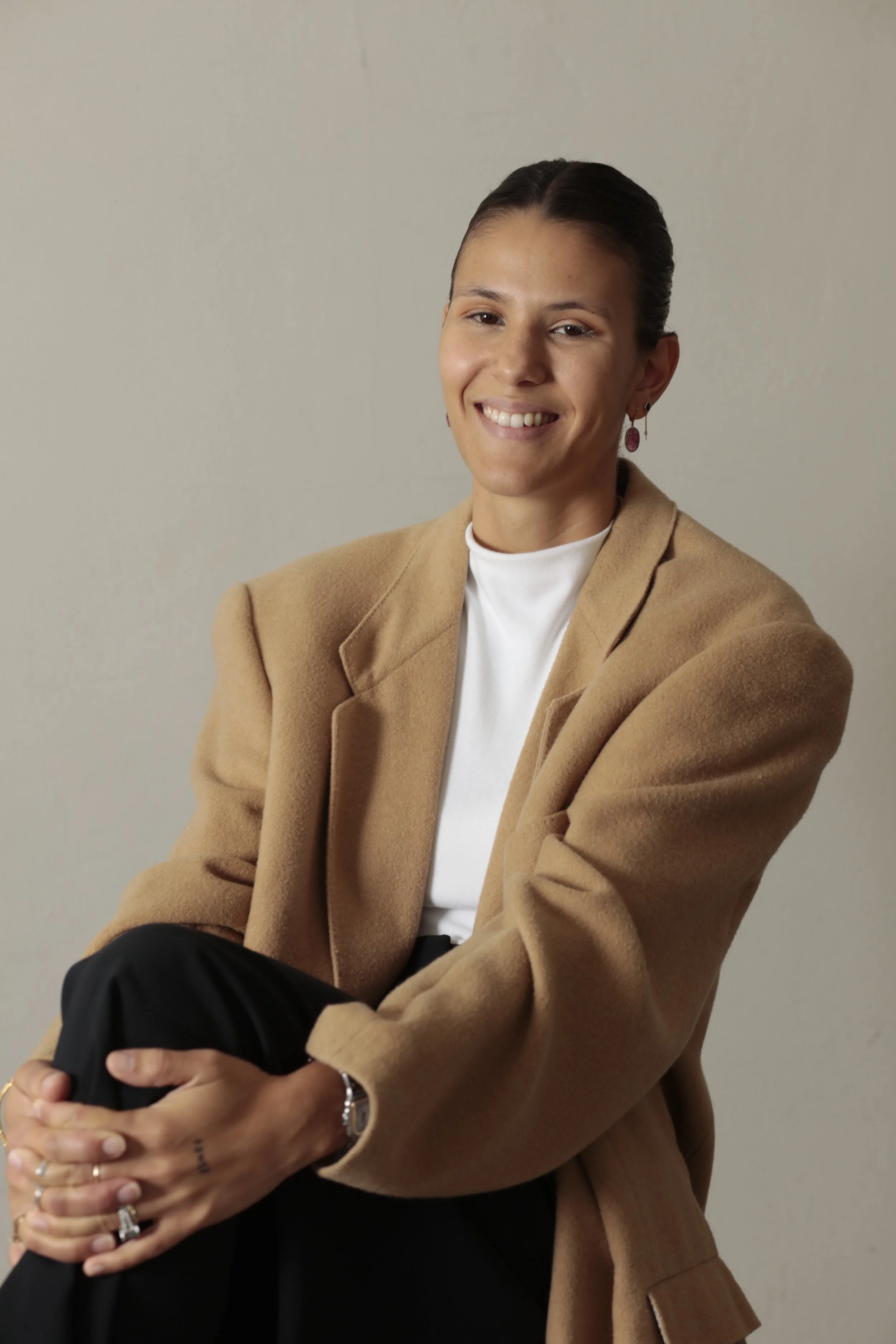 A woman with dark hair tied back, wearing a beige blazer and white t-shirt, smiling and holding someone in her hands, against a plain neutral background.