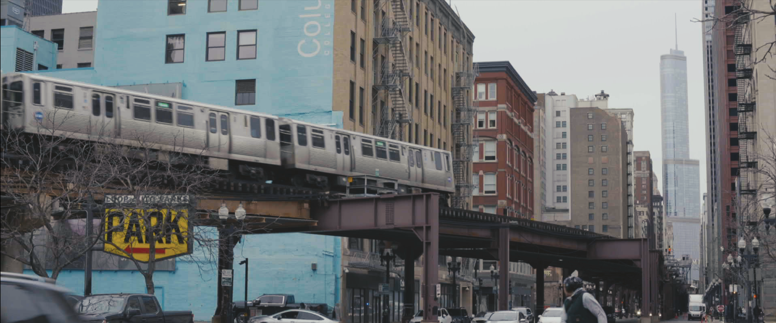 An elevated train traveling through a city street in an urban neighborhood with tall buildings, parking signs, and parked cars.