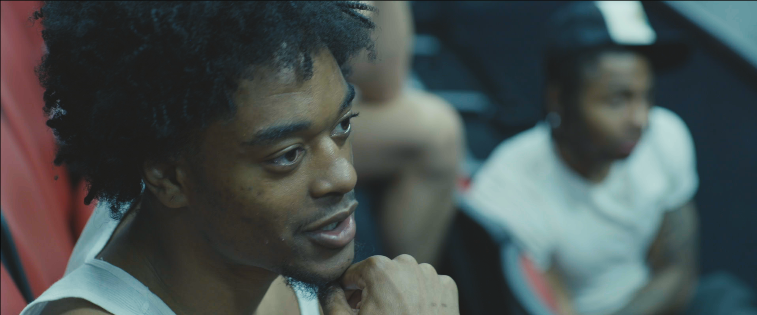Close-up of a young man with curly hair and a light-colored shirt, sitting in a sports arena, attentively watching an event with other spectators in the background.