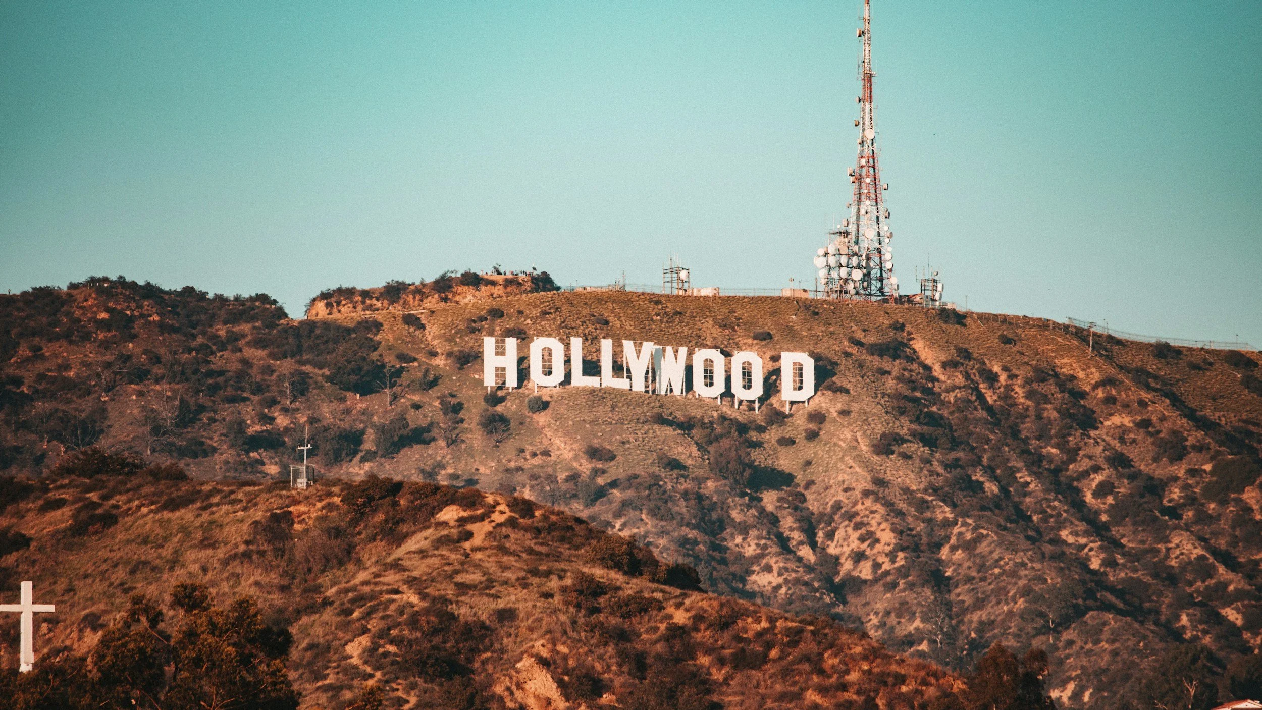 The Hollywood sign on a hillside in Los Angeles, with a communication tower on top and a cross in the foreground.