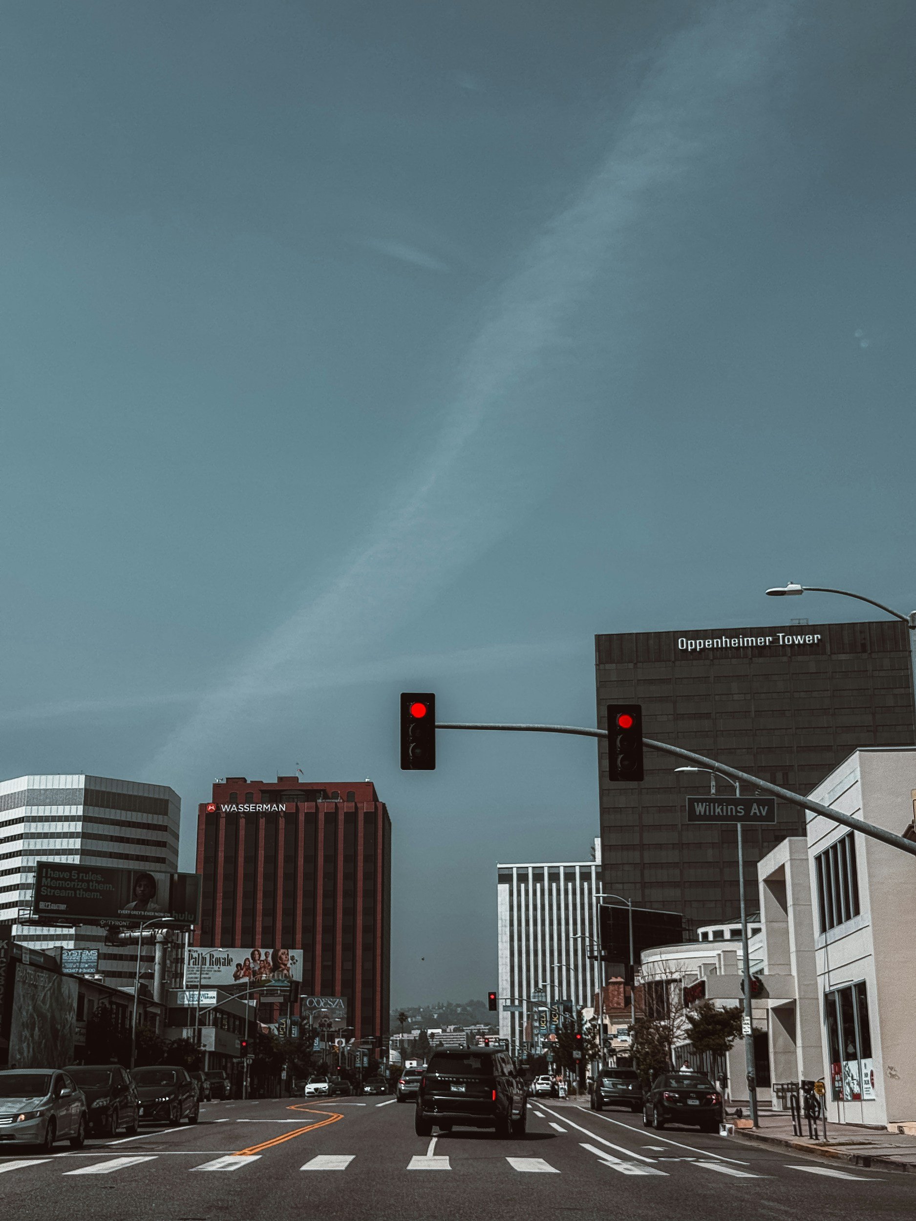 Street scene in downtown Los Angeles with cars at a red traffic light, tall buildings including Oppenheimer Tower and Wasserman, blue sky with wispy clouds, street signs for Wilkins Avenue, and billboard advertisements.