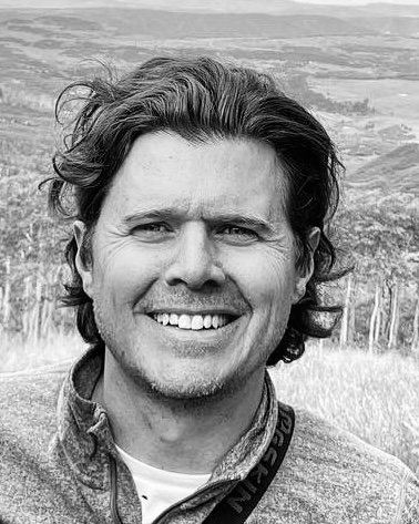 Black and white photo of a smiling man with wavy hair outdoors, with a landscape in the background.