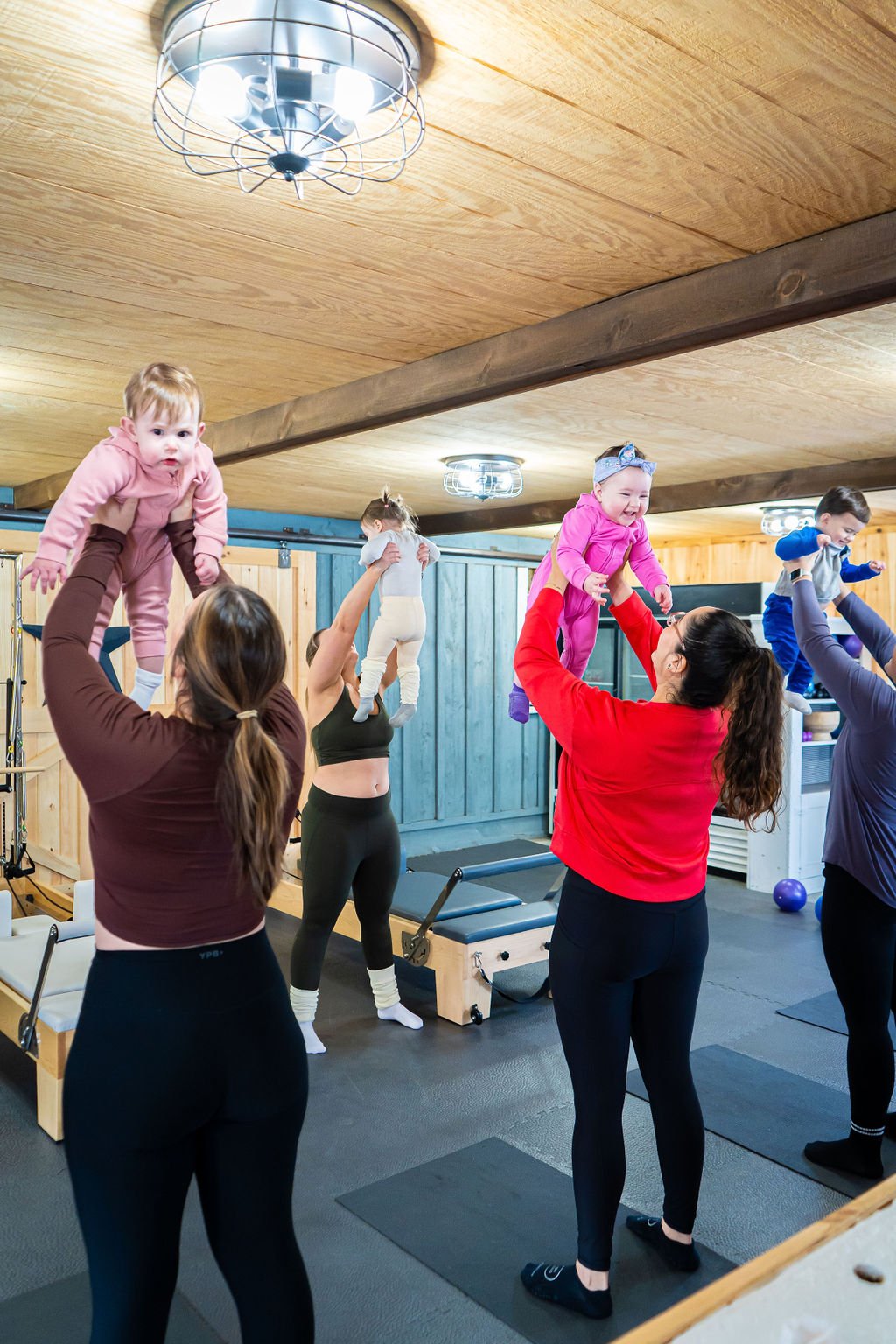 Mothers lifting babies in a fitness class, wooden ceiling, exercise equipment, women wearing activewear, colorful baby outfits.