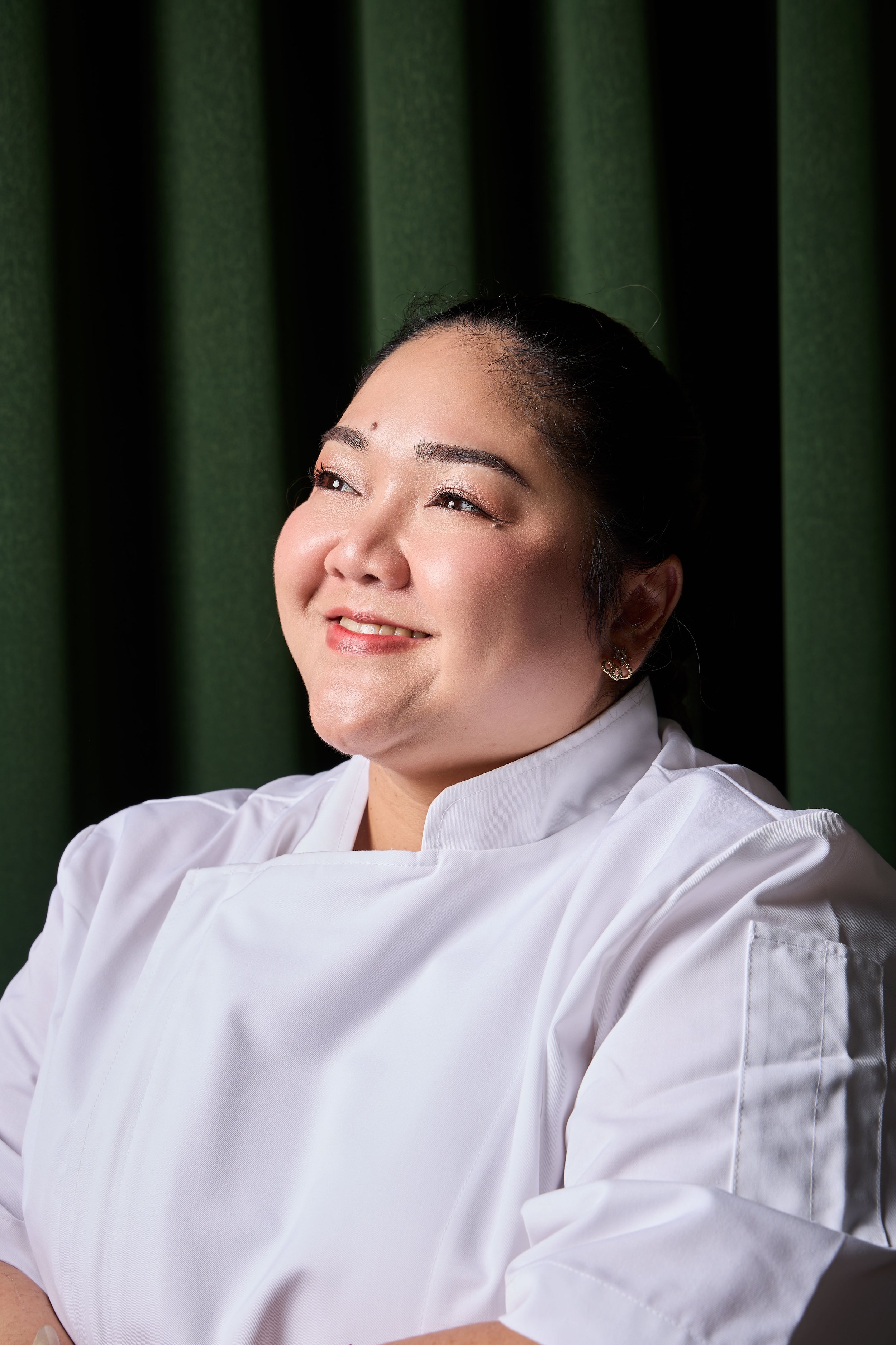 Close-up of a smiling Amita, owner of Khaosan in a white chef's coat with dark hair tied back, standing in front of a green and black background.
