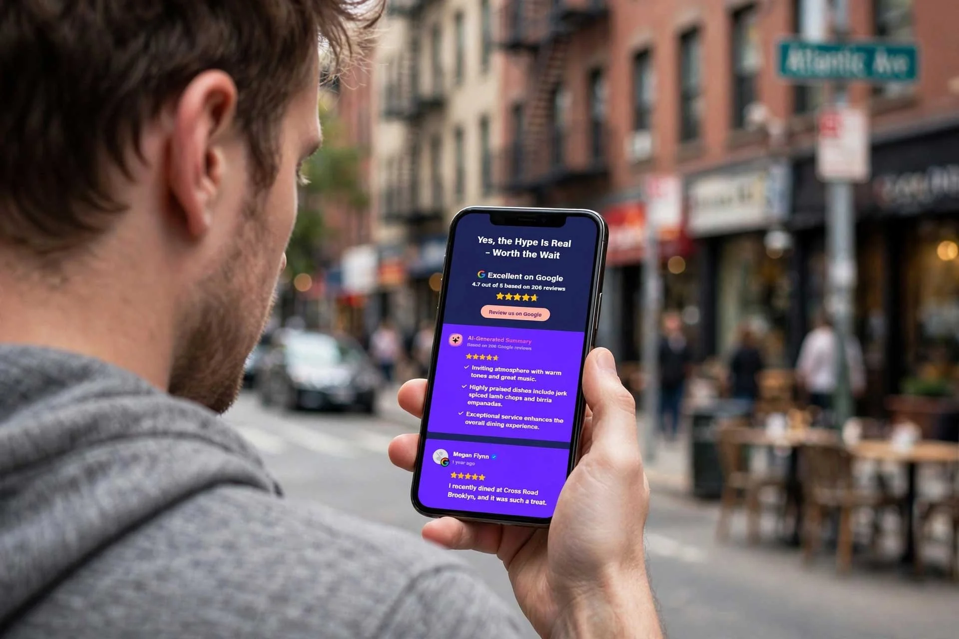 Over-the-shoulder view of a man holding an iPhone displaying a mobile website with google reviews, with a softly blurred Brooklyn street and brownstone buildings in the bright natural daylight background.