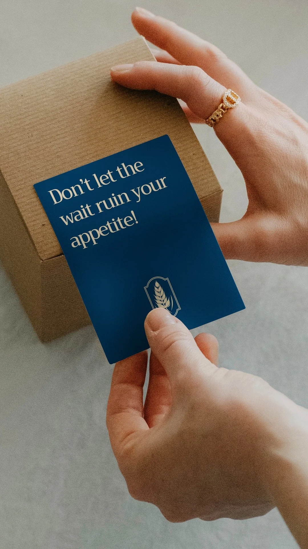 A person holds a blue card with a creative design reading Don't let the wait ruin your appetite over a brown shipping box.