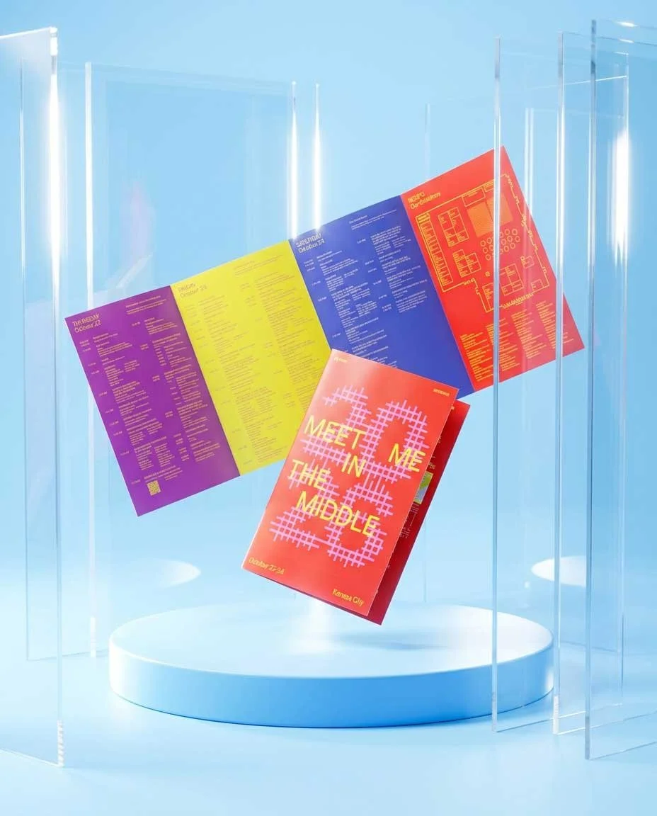 Open colorful brochure and a folded red "MEET ME IN THE MIDDLE" brochure floating above a round white podium, framed by tall clear acrylic panels in a light blue studio.