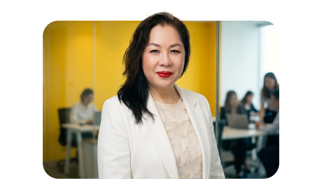 Professional woman with dark hair and red lipstick standing in an office with a yellow wall, with other colleagues working in the background.