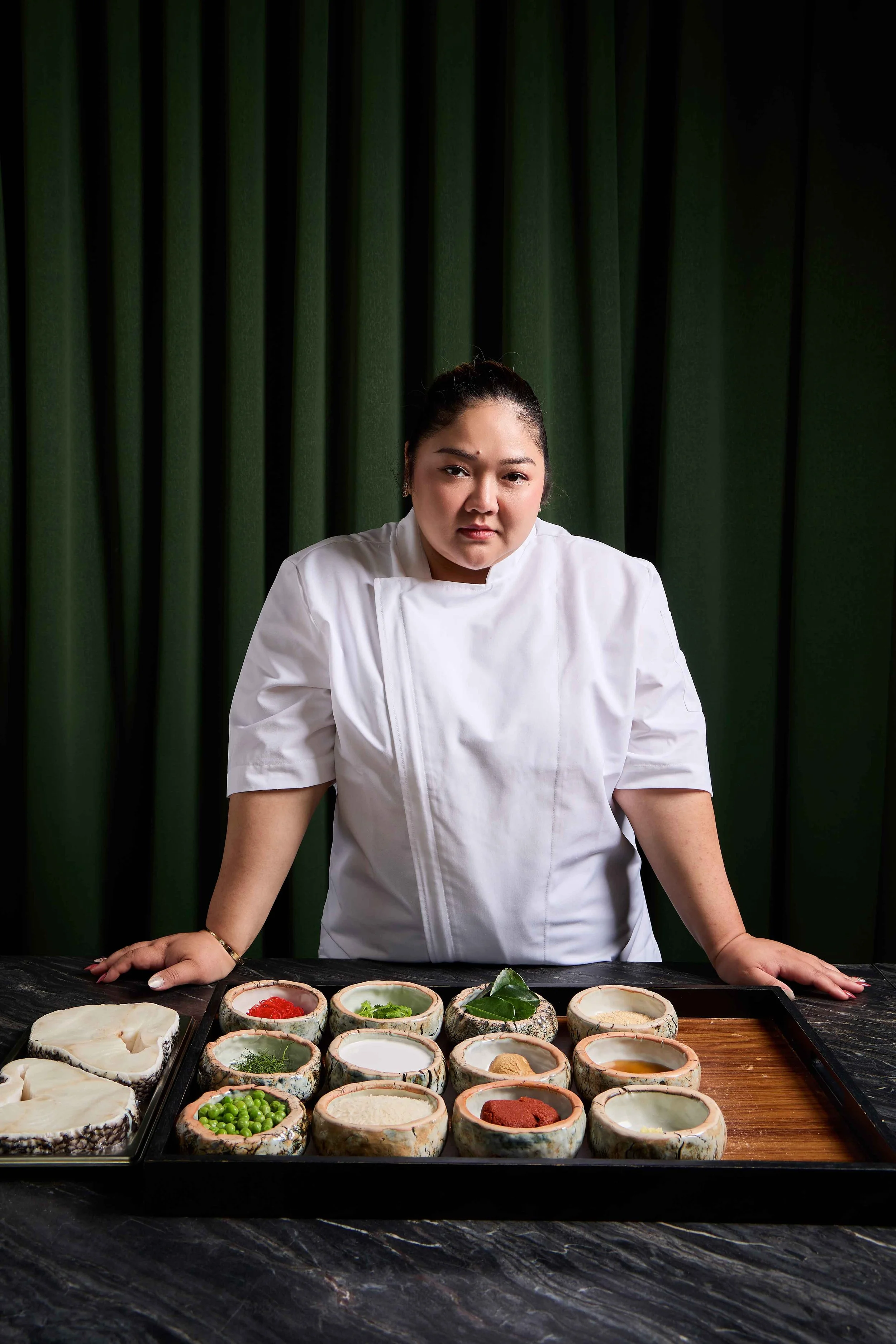 Amita, the owner of Khaosan, in a white uniform, stands behind a table with various bowls of ingredients, including vegetables, herbs, spices, and liquids, on a black marble surface, with green curtains in the background.