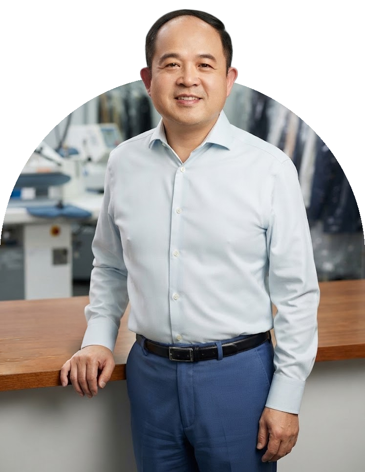 Calvin Jiang, owner of Happy Cleaners in Brooklyn, in a white dress shirt and blue trousers, standing at a wooden counter with a blurred electronics store in the background.
