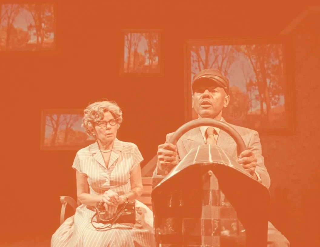 A scene from a museum exhibit or stage show featuring a woman and a man, with the man driving a small, vintage race car and the woman sitting beside him holding a purse, both inside a room with windows showing trees outside.