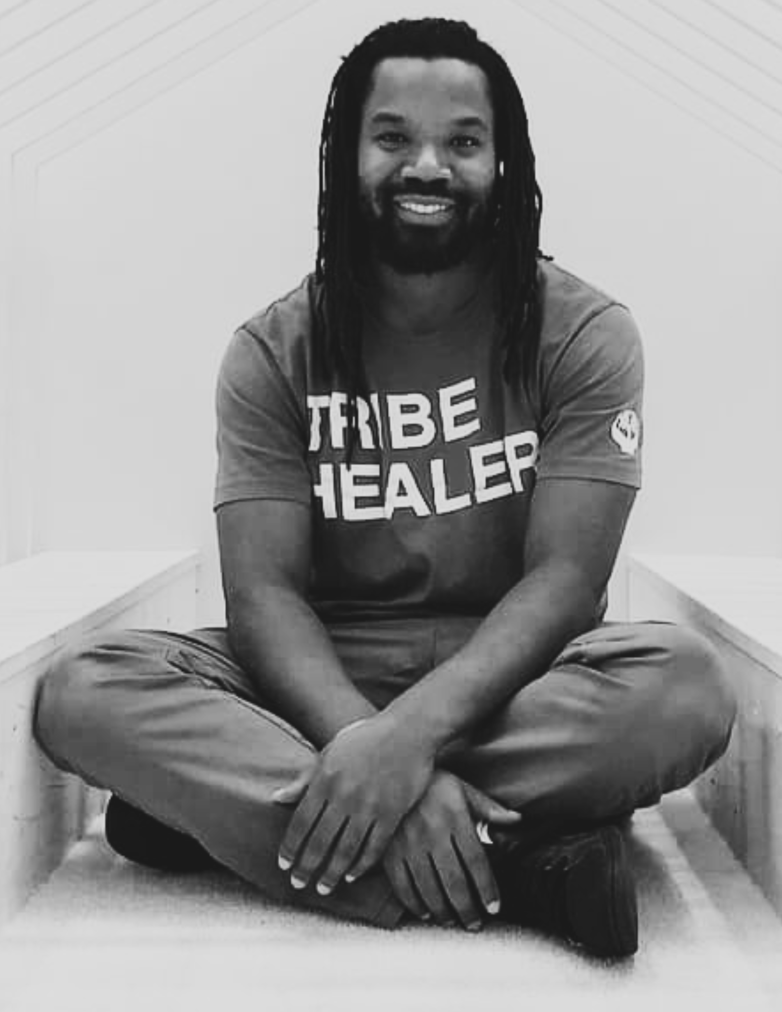 John Johnson of Verbal Gymnastics sitting cross-legged on a platform, smiling at the camera, wearing a T-shirt that says 'TRIBE HEALER'.