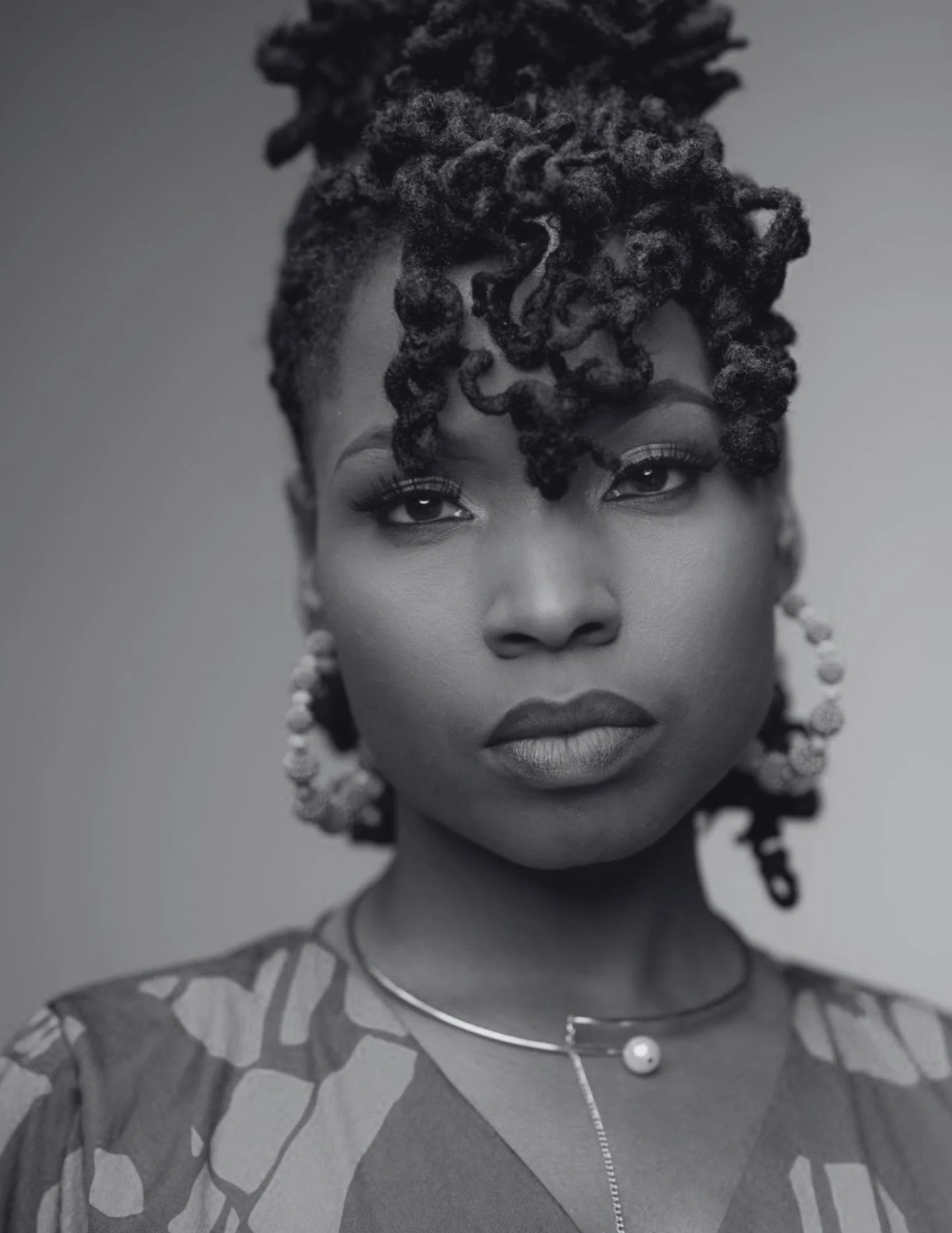 Black and white portrait of a woman with styled dreadlocks, wearing hoop earrings and a necklace with a pearl pendant.