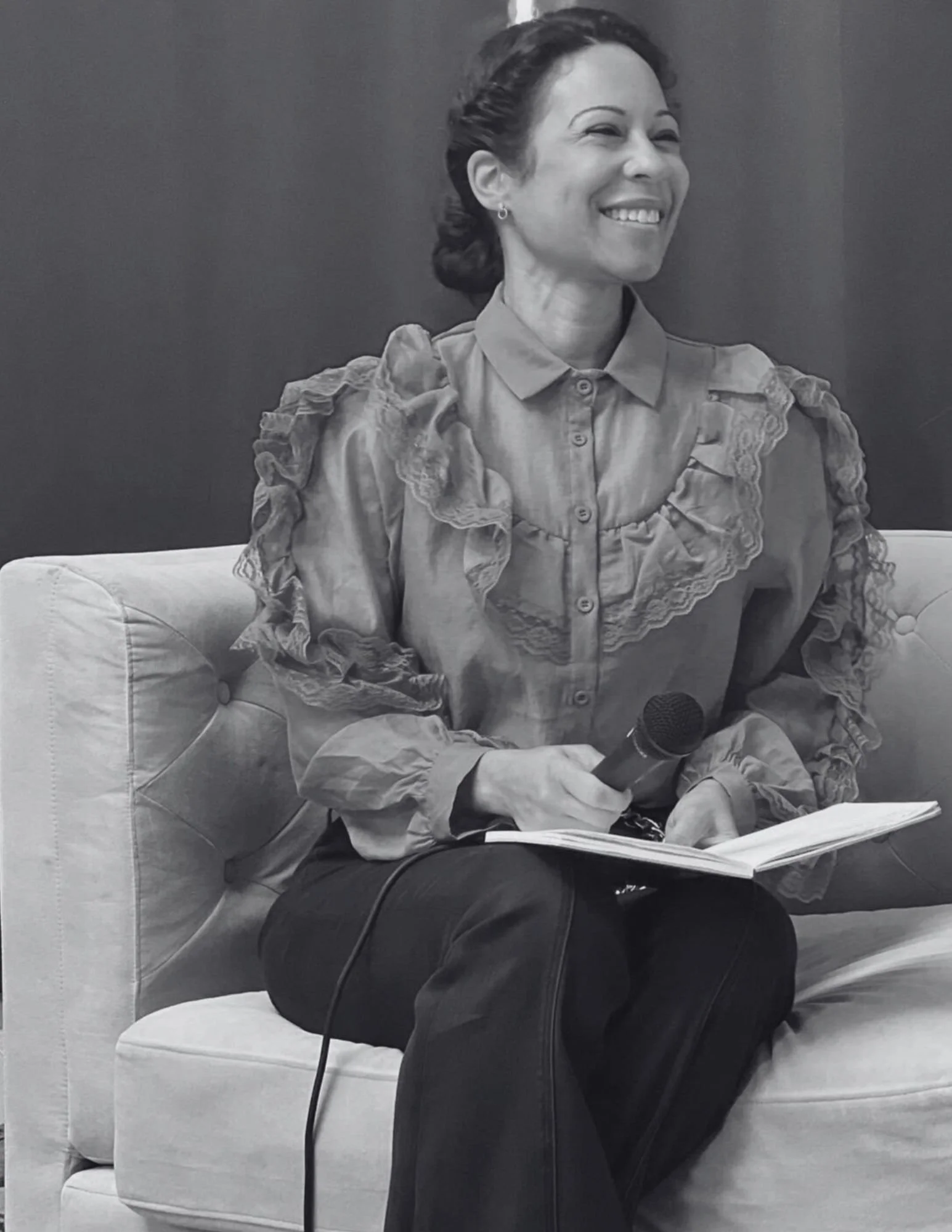 A woman sitting on a sofa, smiling, holding a microphone in her right hand and a notebook in her left hand, against a plain background.