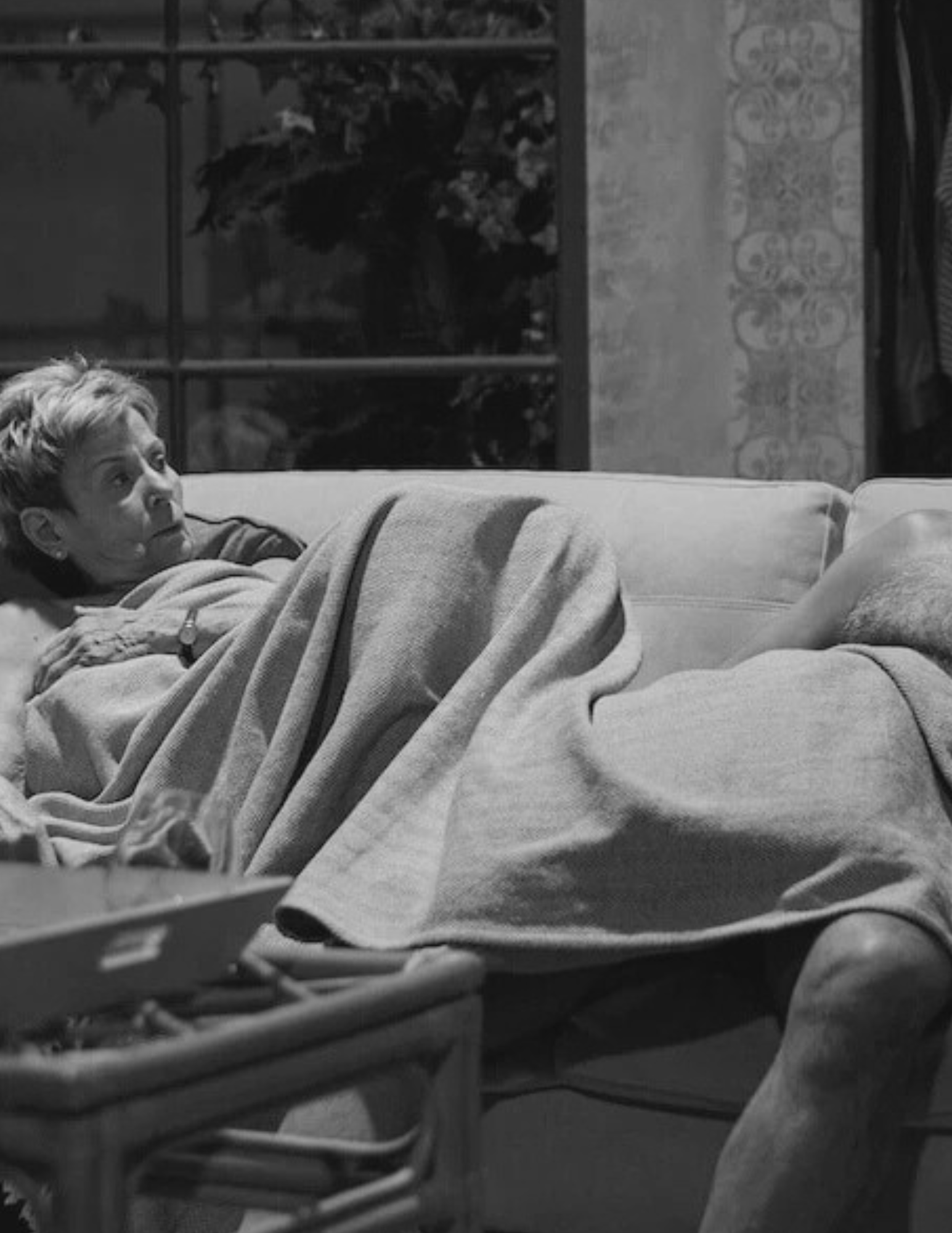 An elderly woman lying on a couch, covered with a blanket, looking pensively to the left. The room has a window with flowers outside and a patterned wallpaper.