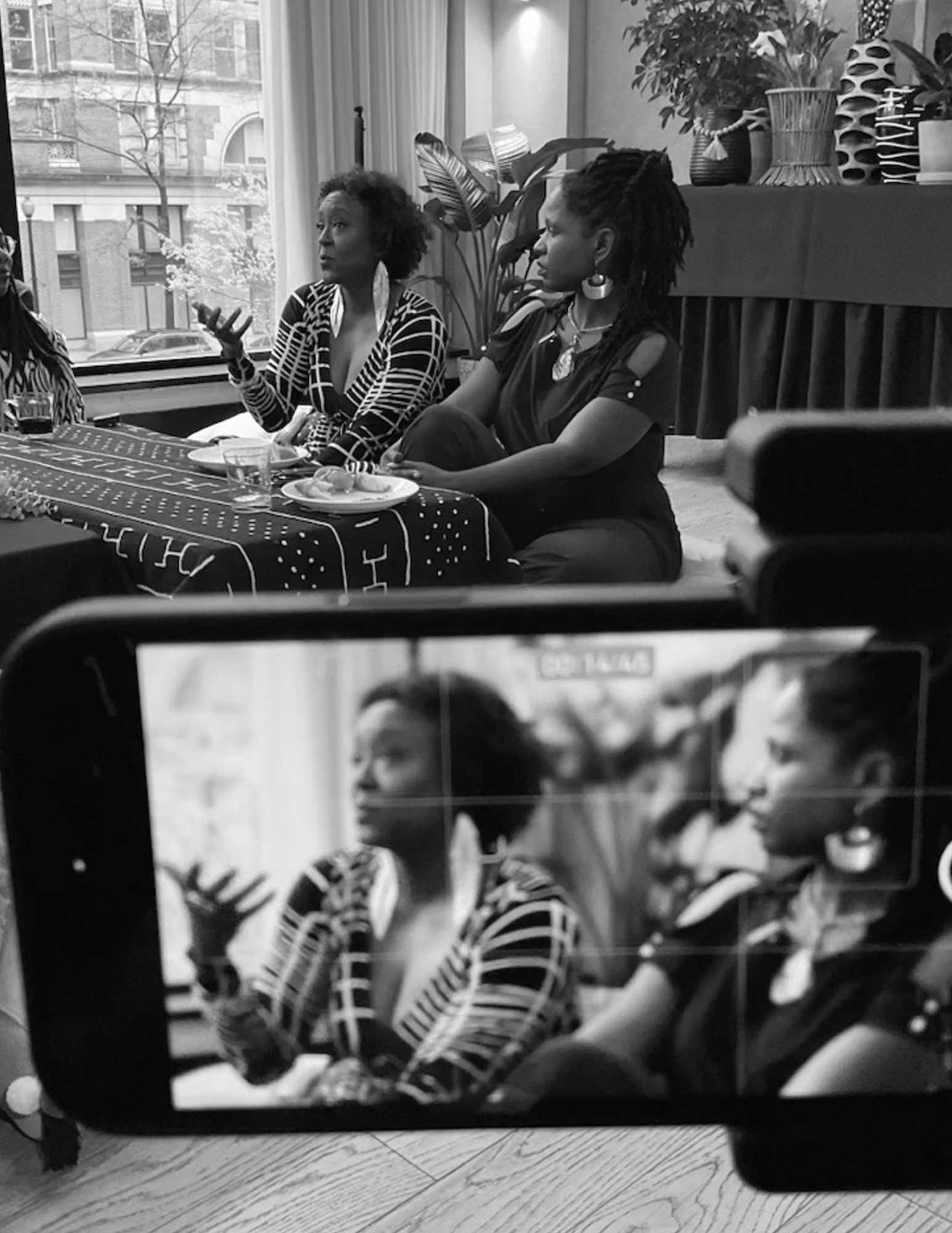 Two women sitting at a table during a discussion or interview, with a camera recording them, in an indoor setting with houseplants and decorative objects in the background.