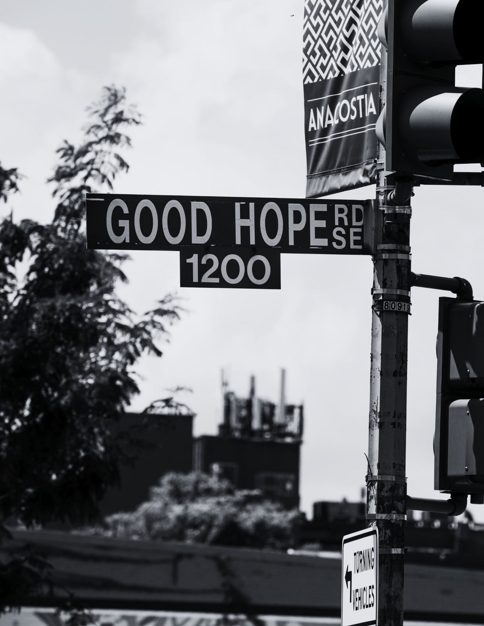 Street sign displaying 'Good Hope Rd SE 1200' at an intersection with traffic lights, a tree, and an advertising banner for Anacostia in the background.