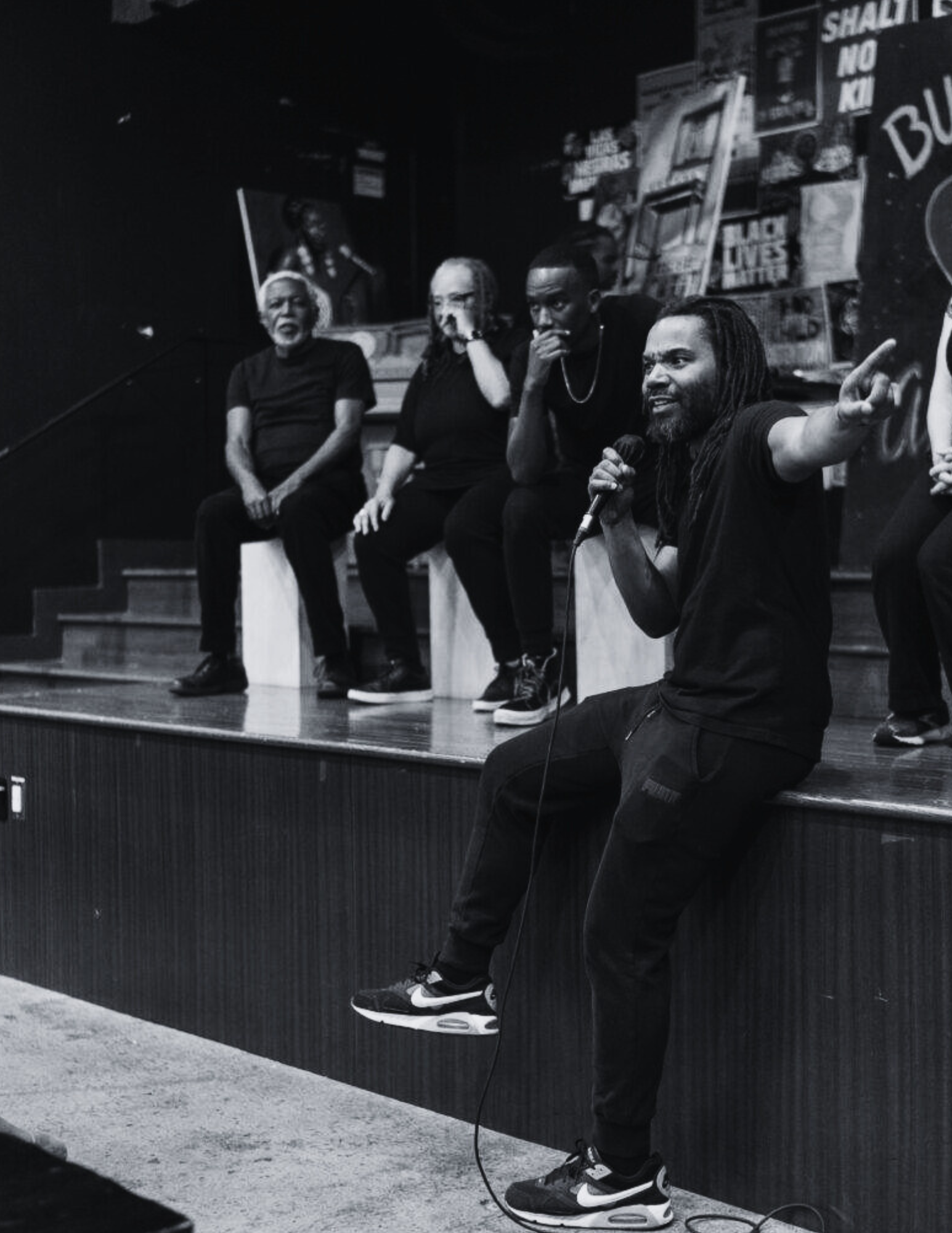 A group of five people sitting on a stage, with one person in the foreground holding a microphone and gesturing, surrounded by posters and artwork on the wall.