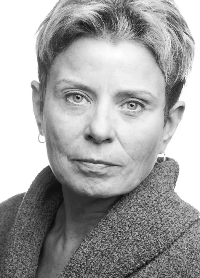 Black and white portrait of a woman with short hair, wearing earrings and a textured coat, looking directly at the camera with a serious expression.