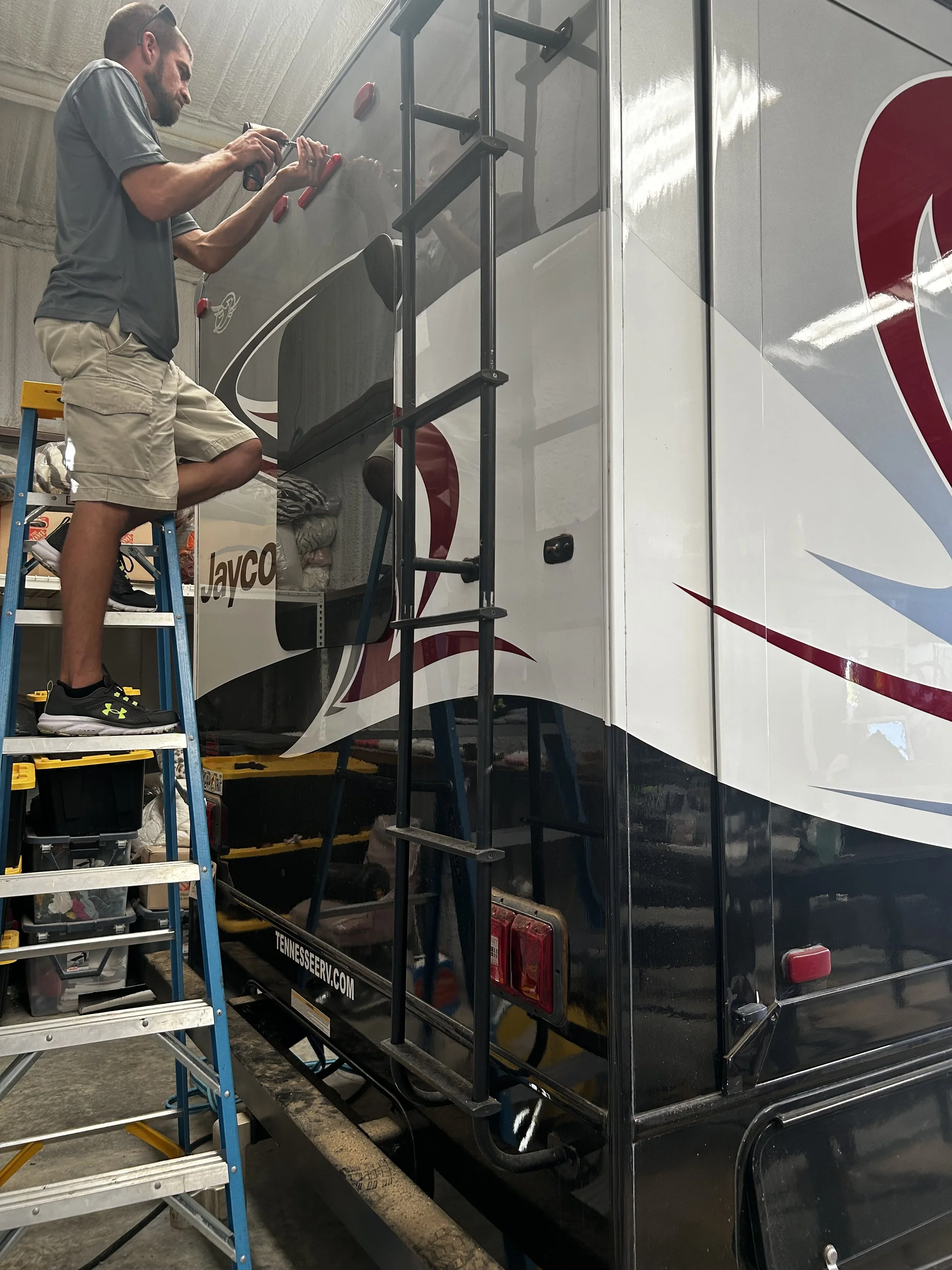 A man standing on a step ladder, polishing the exterior of a large black and white RV with red accents inside a workshop or garage.