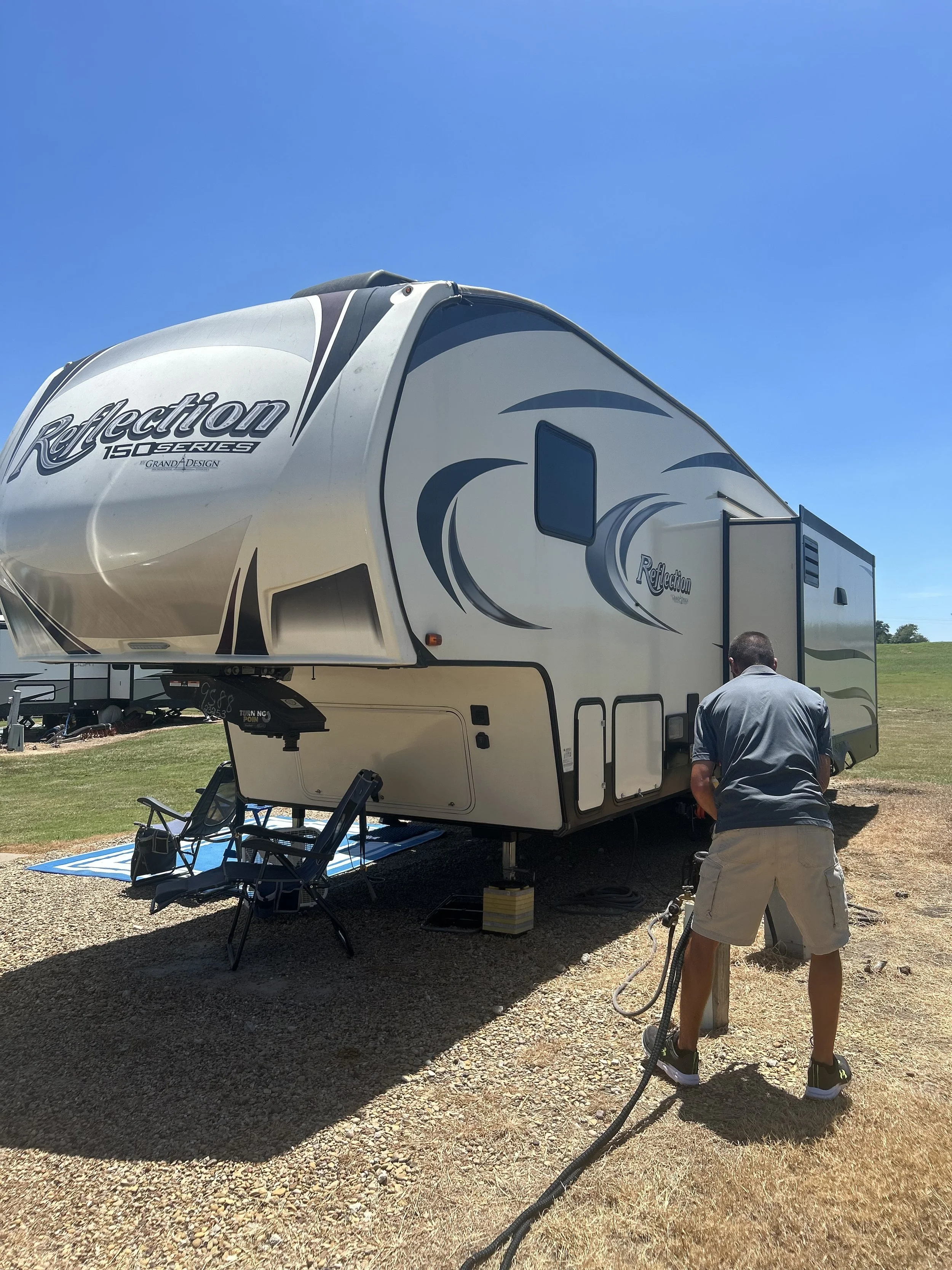 A man is cleaning or inspecting a large Reflection 150Q Series fifth wheel RV trailer on a gravel surface under a clear blue sky. The RV is cream and brown with decorative swirls, and outdoor chairs and a blue and white striped rug are placed underne
