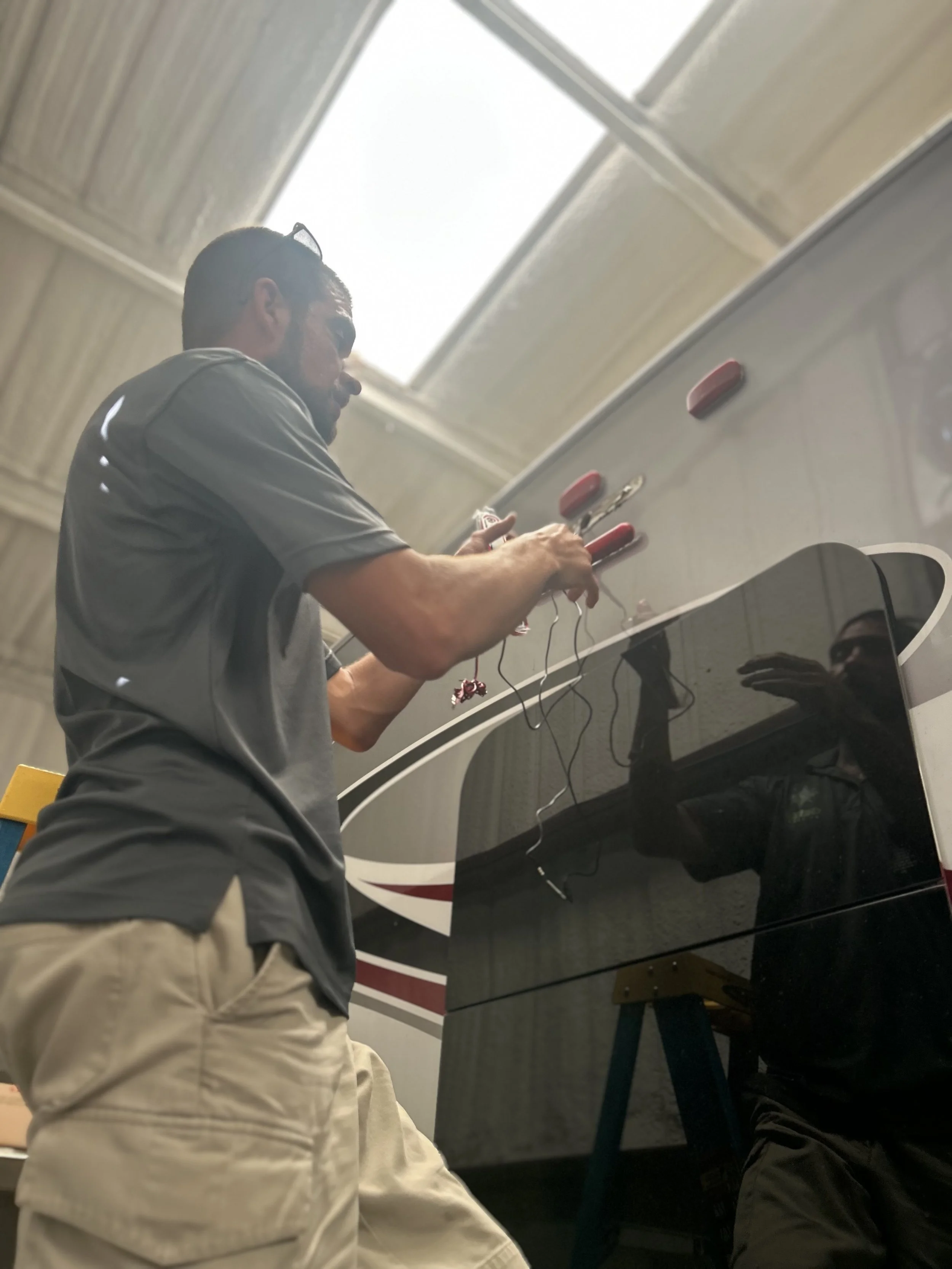 A man working on wiring near the top of a black recreational vehicle or bus in an indoor space with a high ceiling.